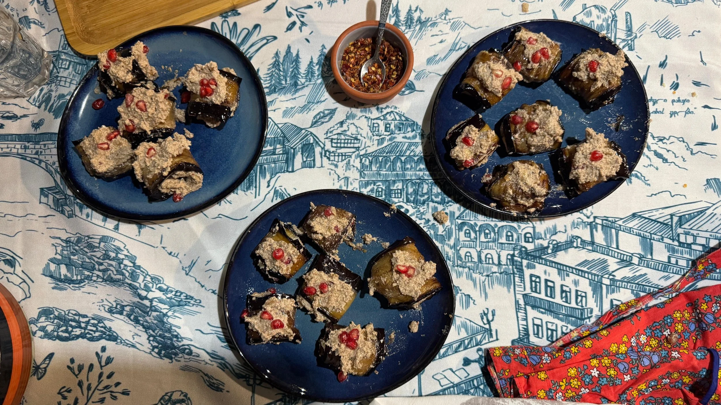 Overhead view of nigvziani badrijani (Georgian eggplant rolls with walnut paste) arranged on three blue plates, garnished with pomegranate seeds, served on a patterned tablecloth