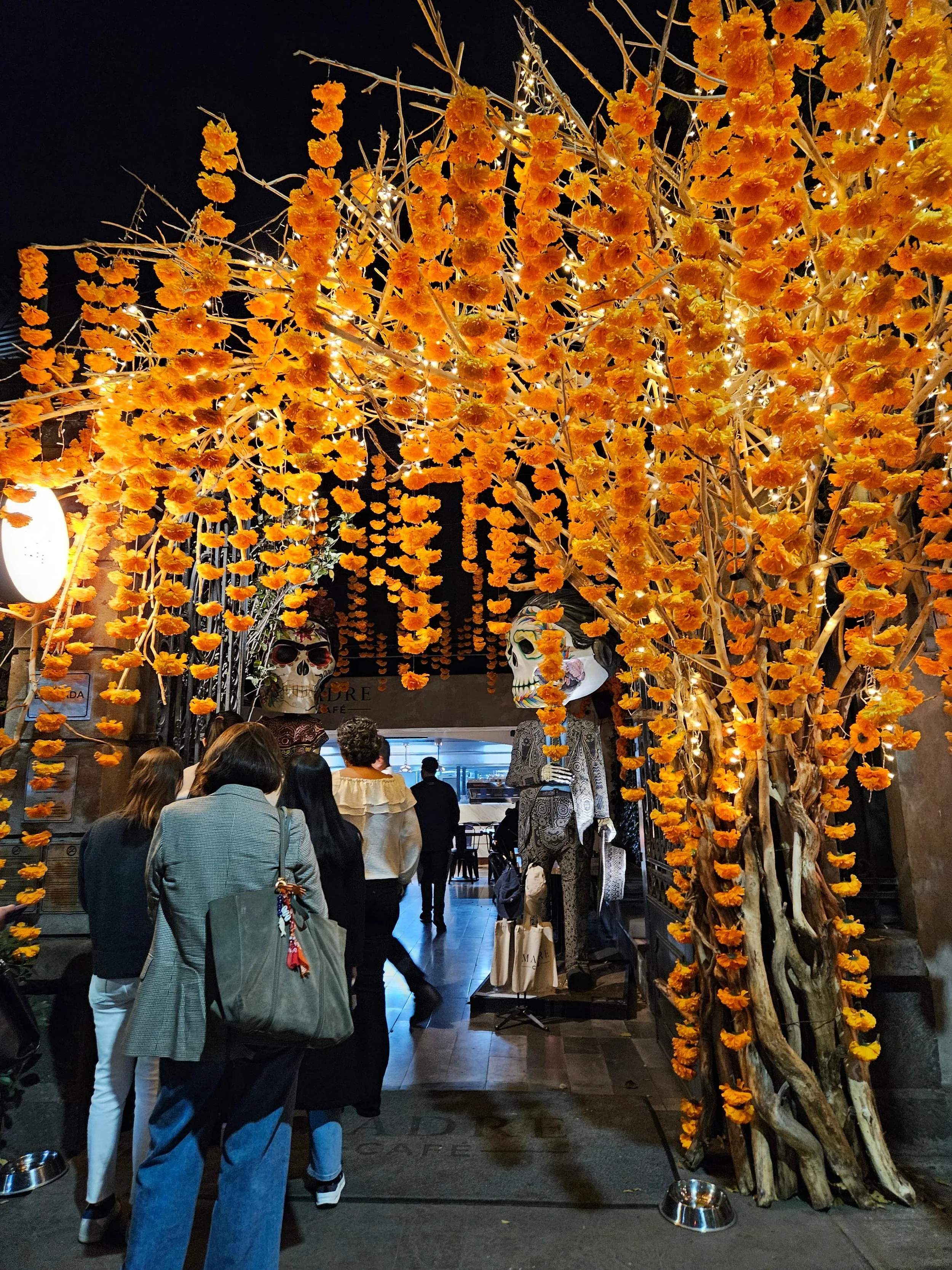 Elaborate Day of the Dead (Día de los Muertos) altar entrance decorated with cascading orange marigold flowers draped over branches, with skeleton figures visible beneath the floral archway and visitors walking through.