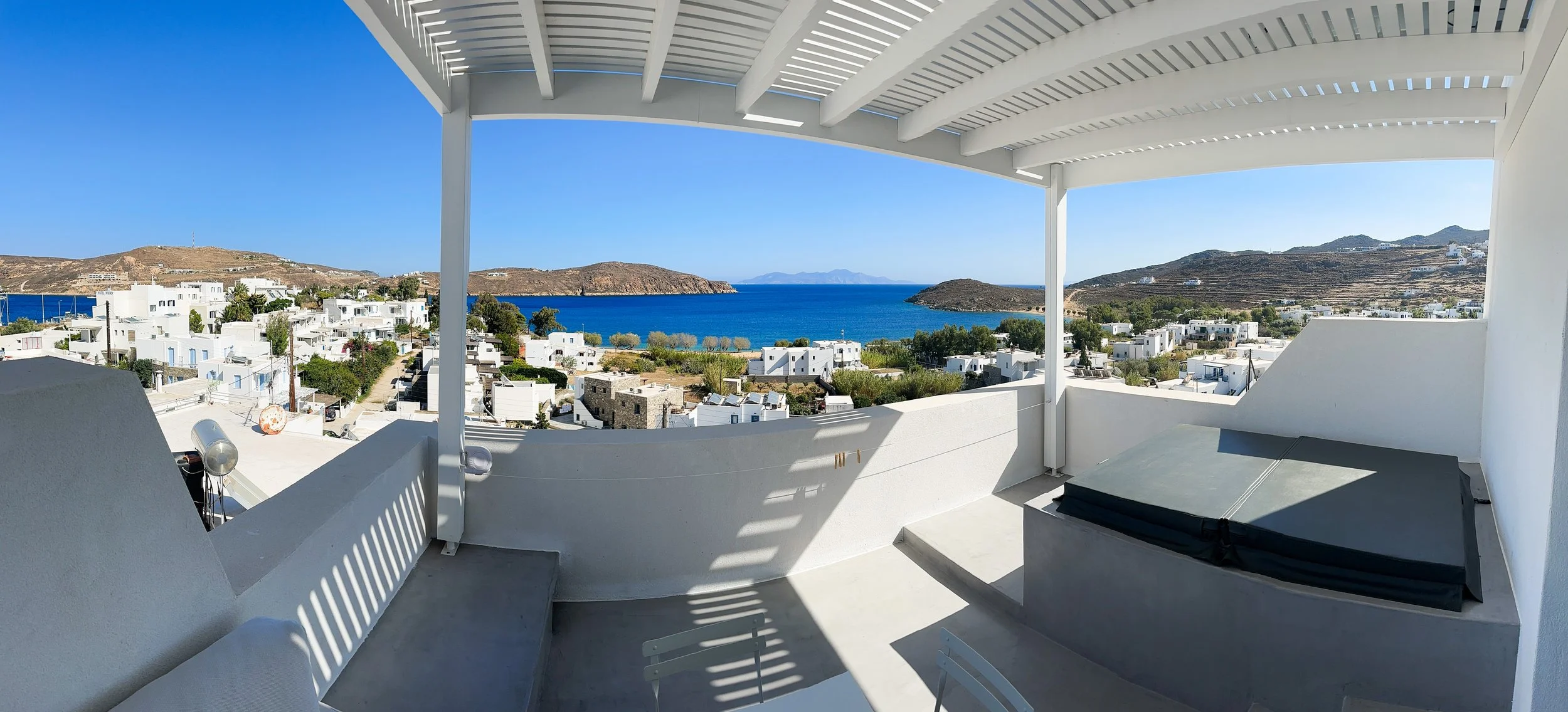 A wide-angle view from a white terrace with a pergola, overlooking the Aegean Sea, surrounding islands, and the white Cycladic houses of Serifos nestled on the hillside.  Copy Retry