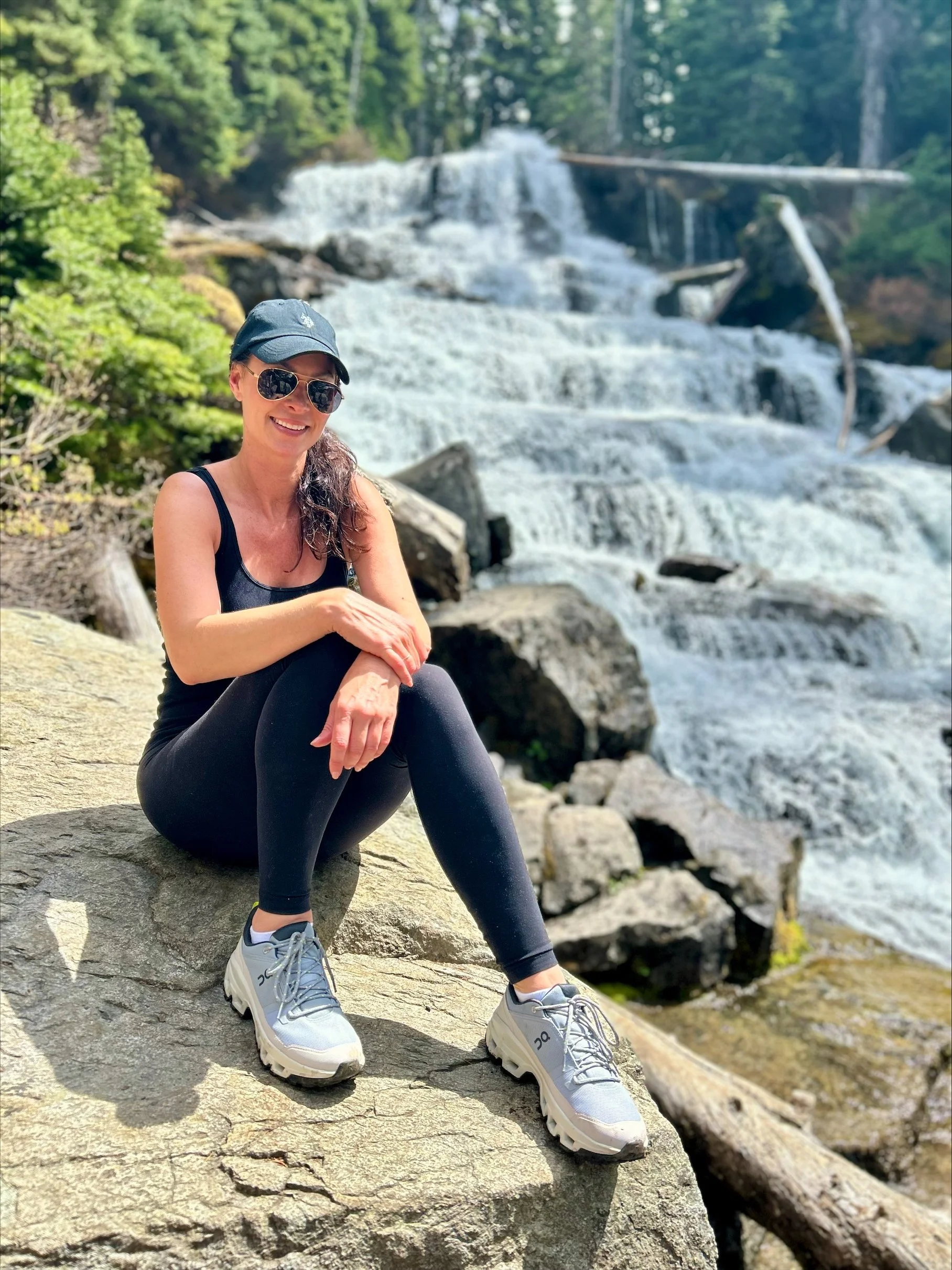 A woman wearing a black baseball cap, sunglasses, black athletic wear, and gray hiking boots sits on stone steps beside a multi-tiered waterfall cascading down rocky terrain.
