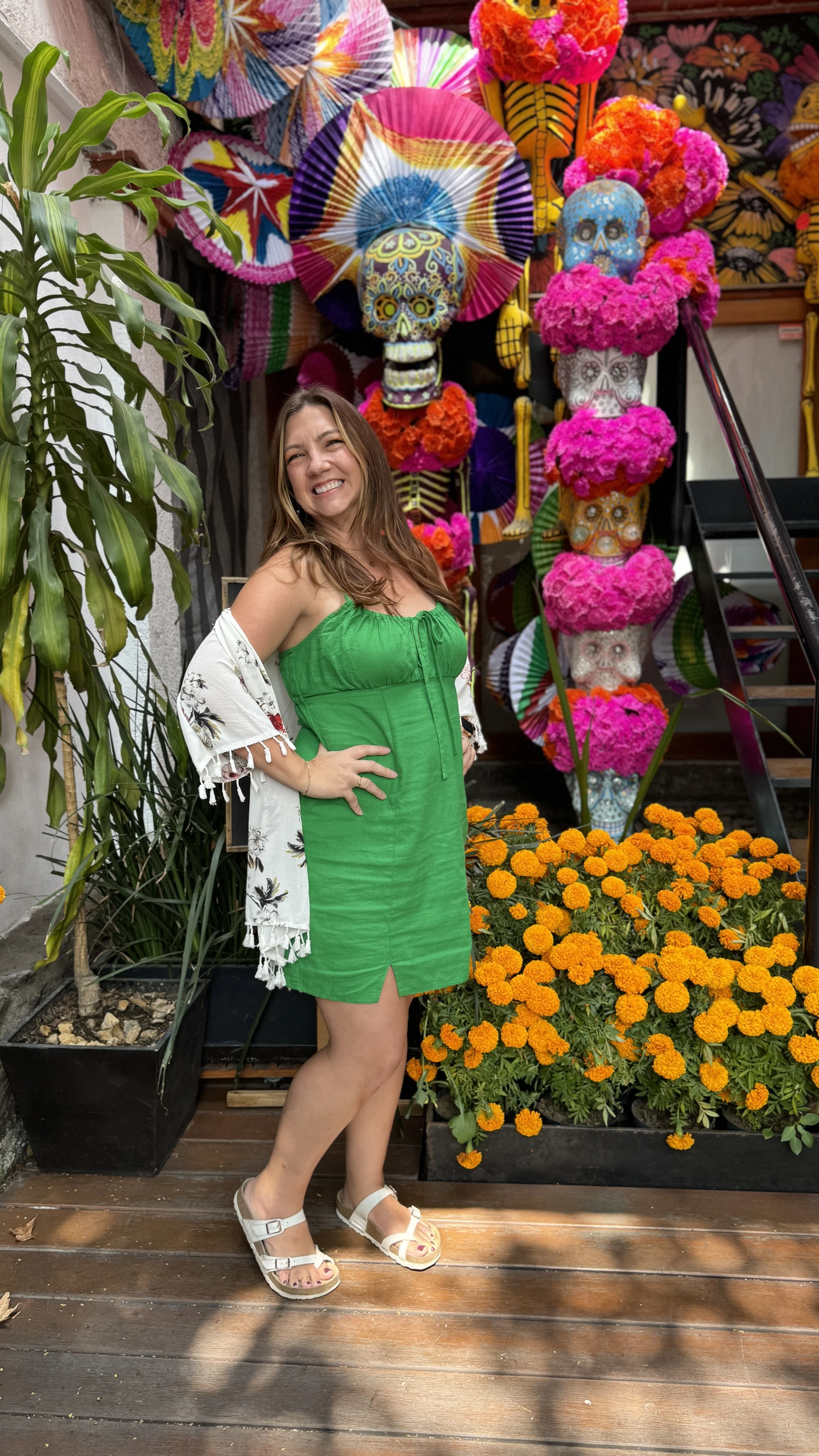 Woman in green dress smiling in front of colorful Day of the Dead decorations including decorated skulls with bright pink and orange marigold flowers in Mexico City