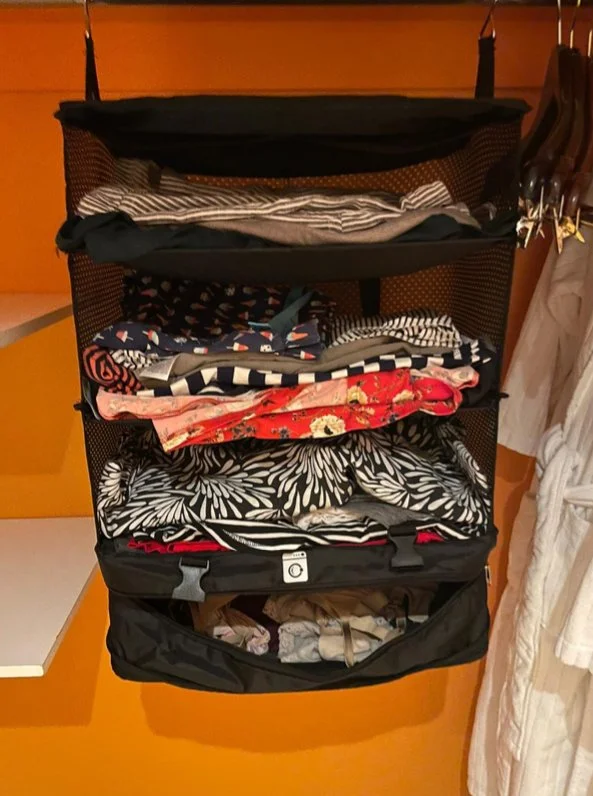 Black hanging travel organizer with multiple compartments filled with folded clothes, hanging in a hotel closet with orange walls.