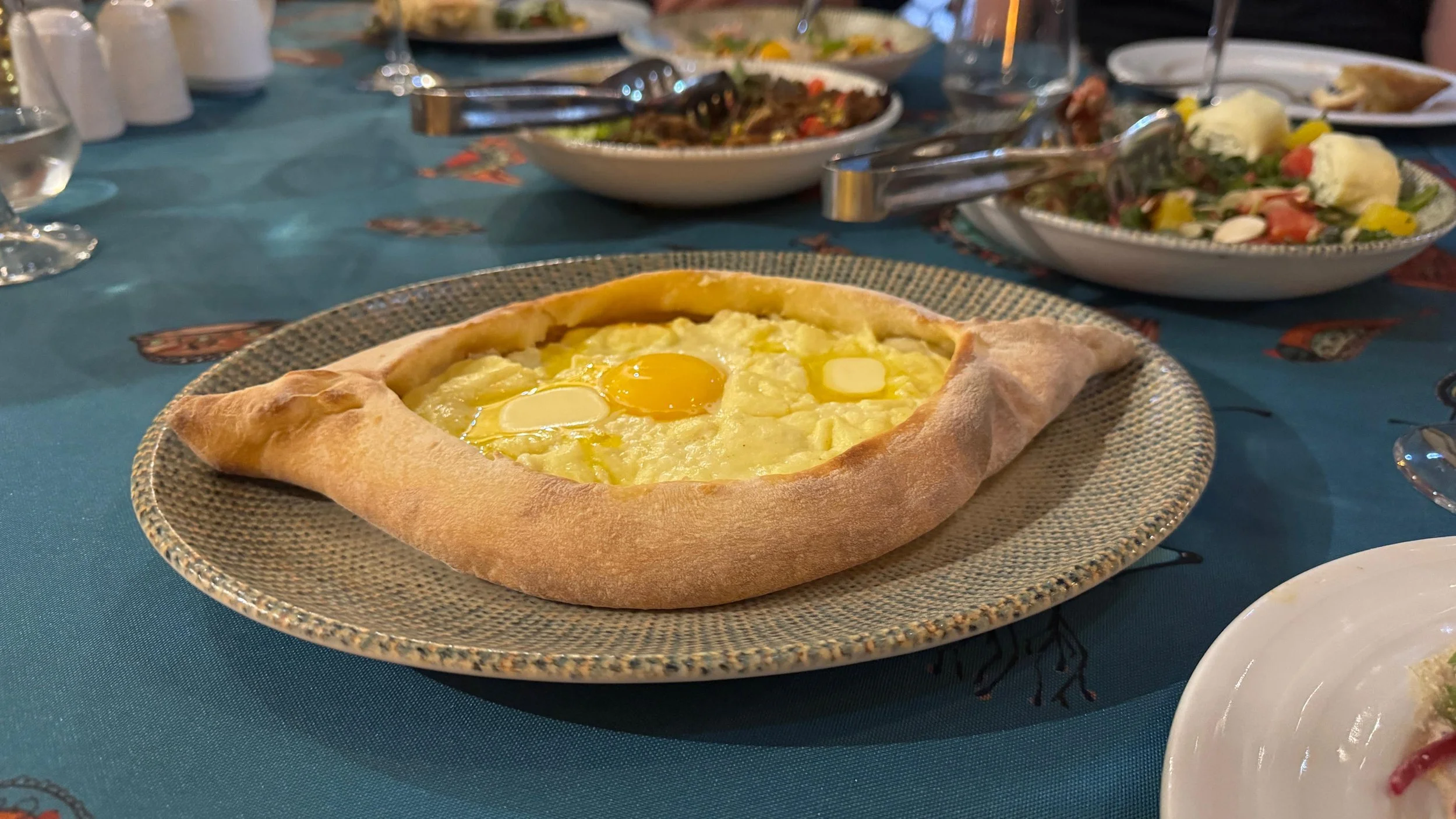 Boat-shaped Adjarian khachapuri on a decorative plate at a Georgian restaurant table, filled with melted cheese, raw egg yolk, and butter in the center