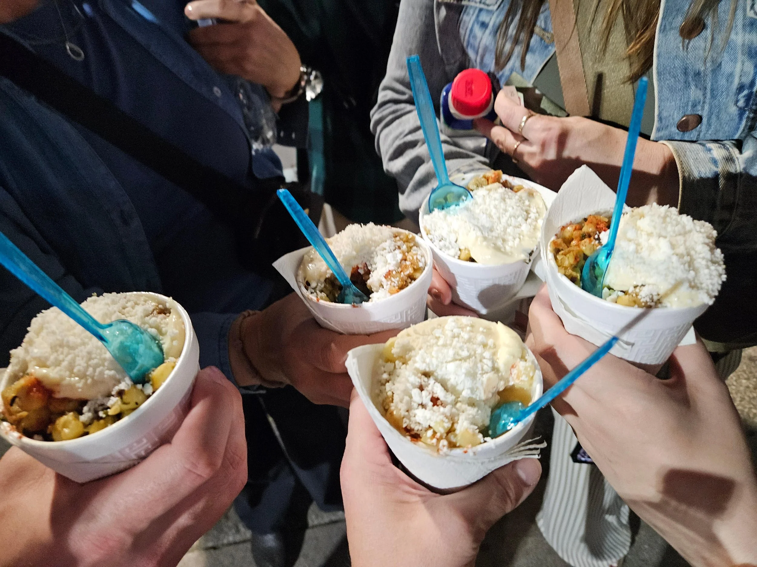 Group of travelers holding white cups filled with street food topped with rice and blue spoons, gathered together in what appears to be a Mexican market or street vendor setting.