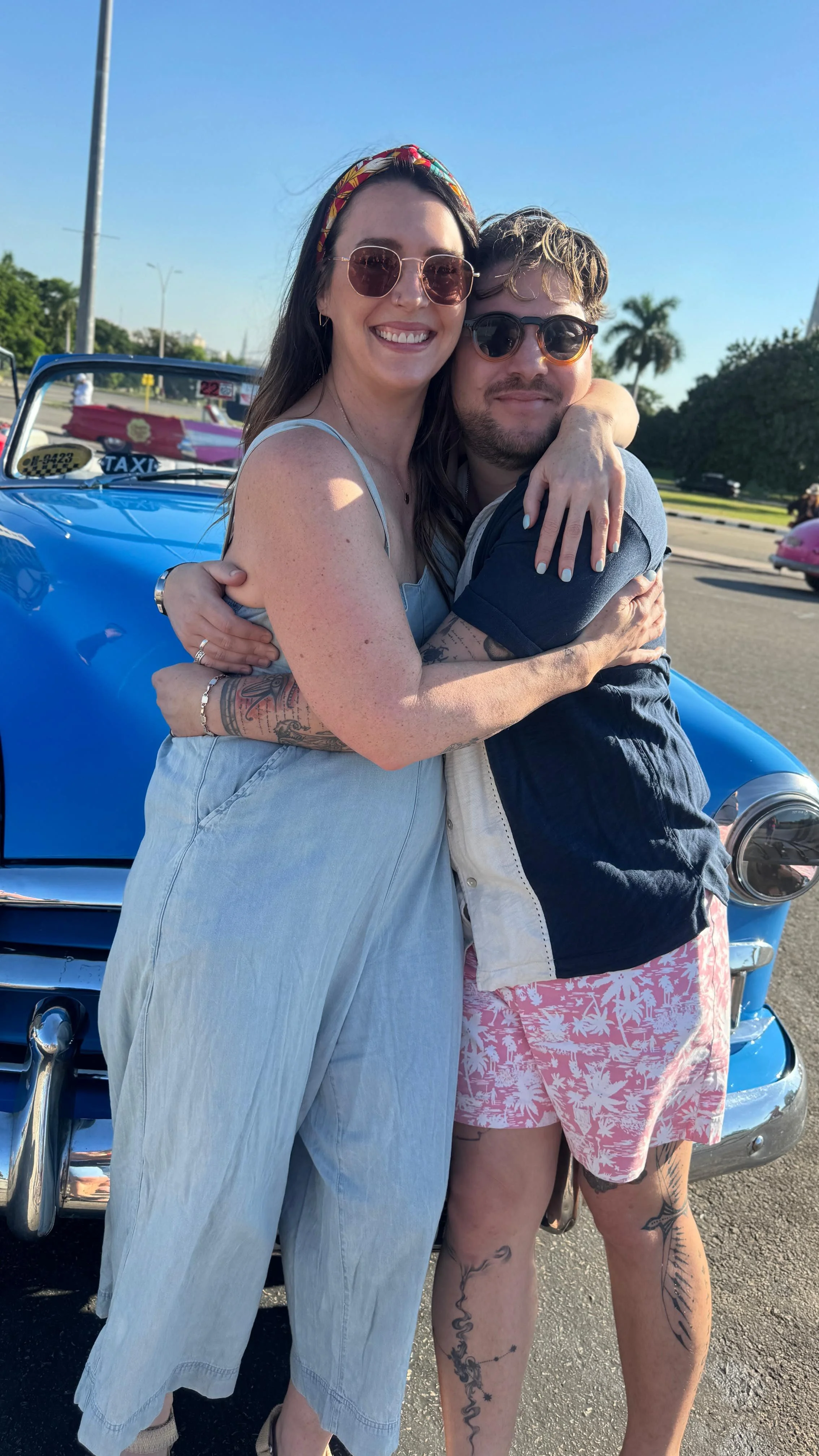 Laura Ericson and guide Andrés hugging in front of a blue classic car taxi in Havana, Cuba.