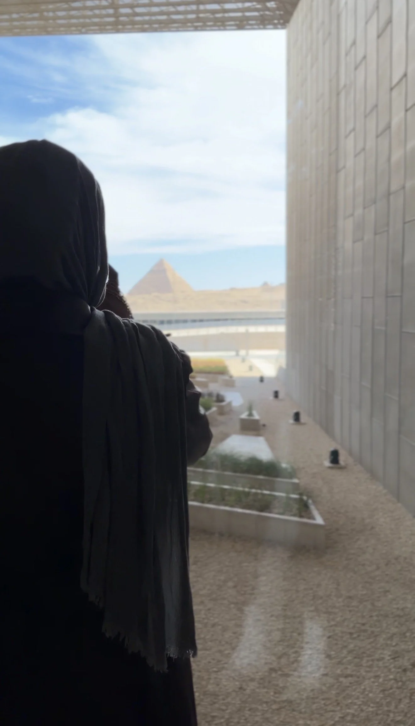 Person wearing a headscarf looking out a window at a pyramid in the distance, outside of a building with brick walls.