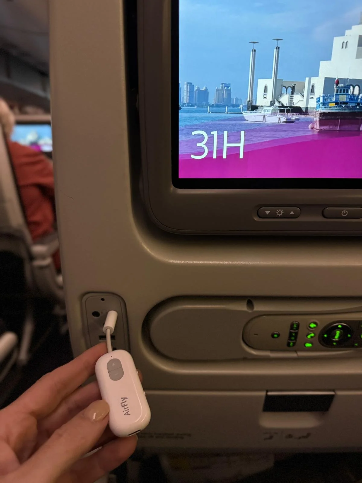 Hand holding a white AirFly Bluetooth adapter plugged into an airplane seatback headphone jack below the in-flight entertainment screen