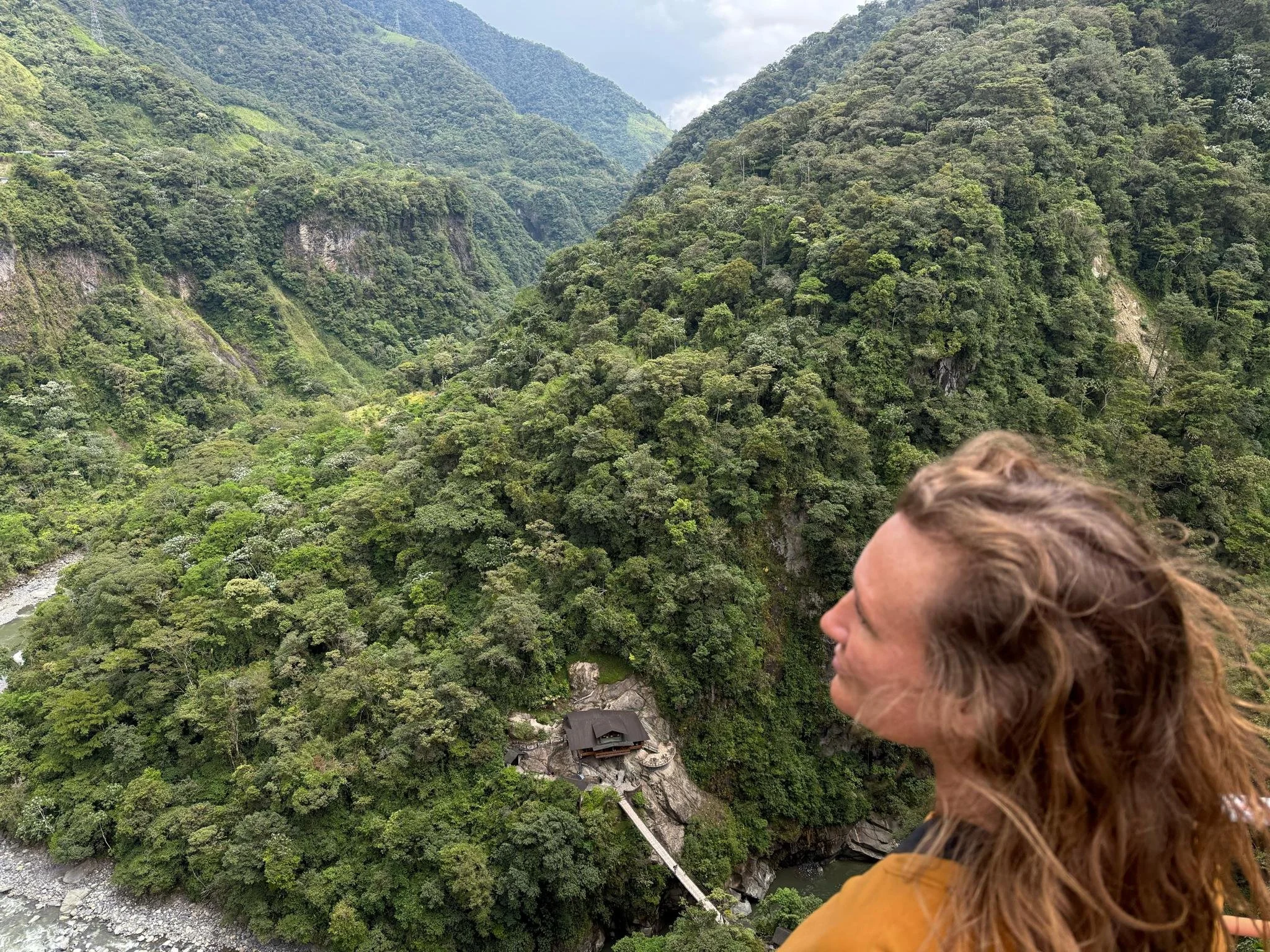 Woman looking out over a dramatic jungle canyon with a small lodge nestled at the base of the cliffs in Baños, Ecuador