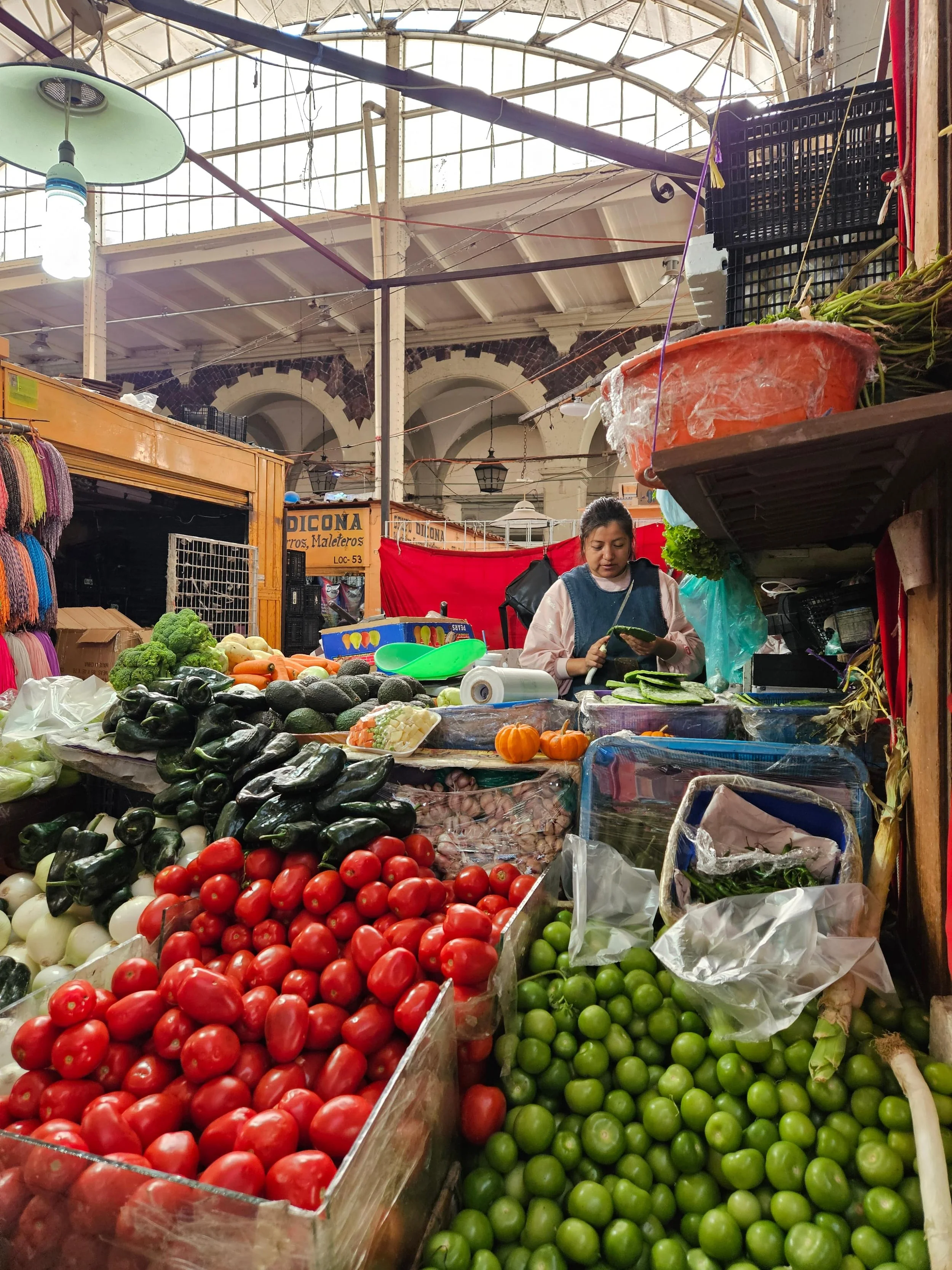 Vendor at a traditional Mexican market stall surrounded by fresh produce including tomatoes, limes, avocados, and various vegetables under a covered market structure with natural lighting.