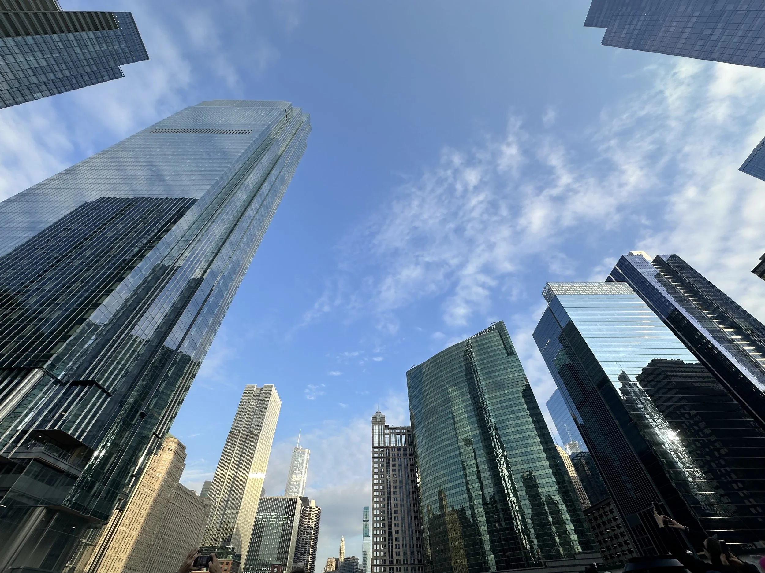 An upward-looking perspective of towering glass skyscrapers framing a bright blue sky with wispy clouds in a dense urban downtown.