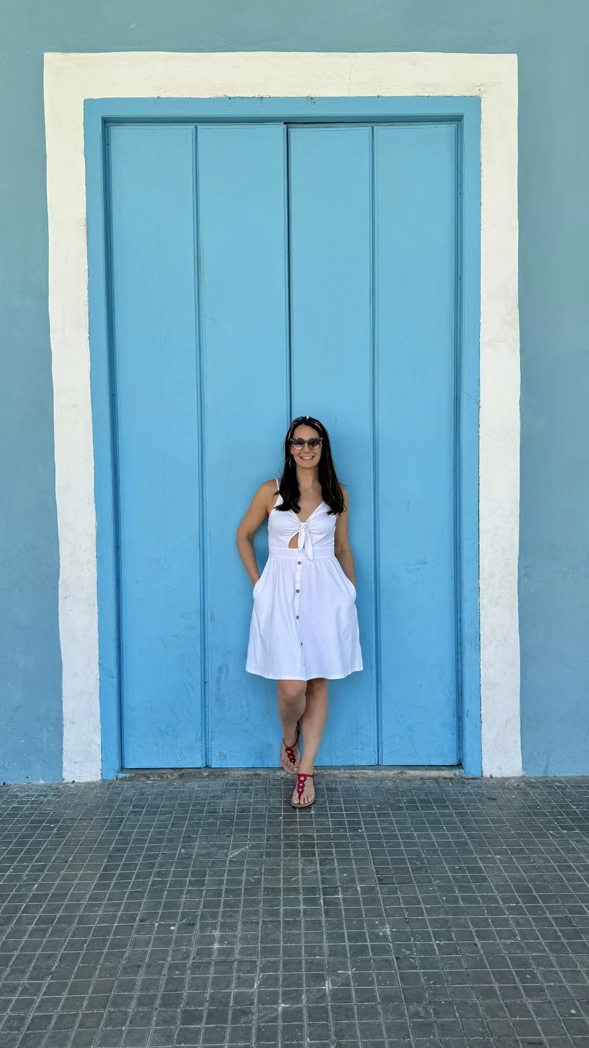 Woman in white button-front dress and red sandals posing in front of bright turquoise blue wooden doors with white trim on a tiled courtyard floor.