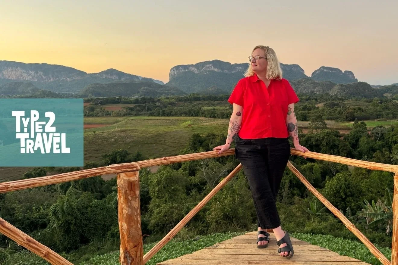 Mel Ripp in a bright red shirt and black pants standing on a wooden viewing platform at sunset, with lush green landscape, turquoise water, and dramatic mountains in the background, Type 2 Travel podcast logo in corner