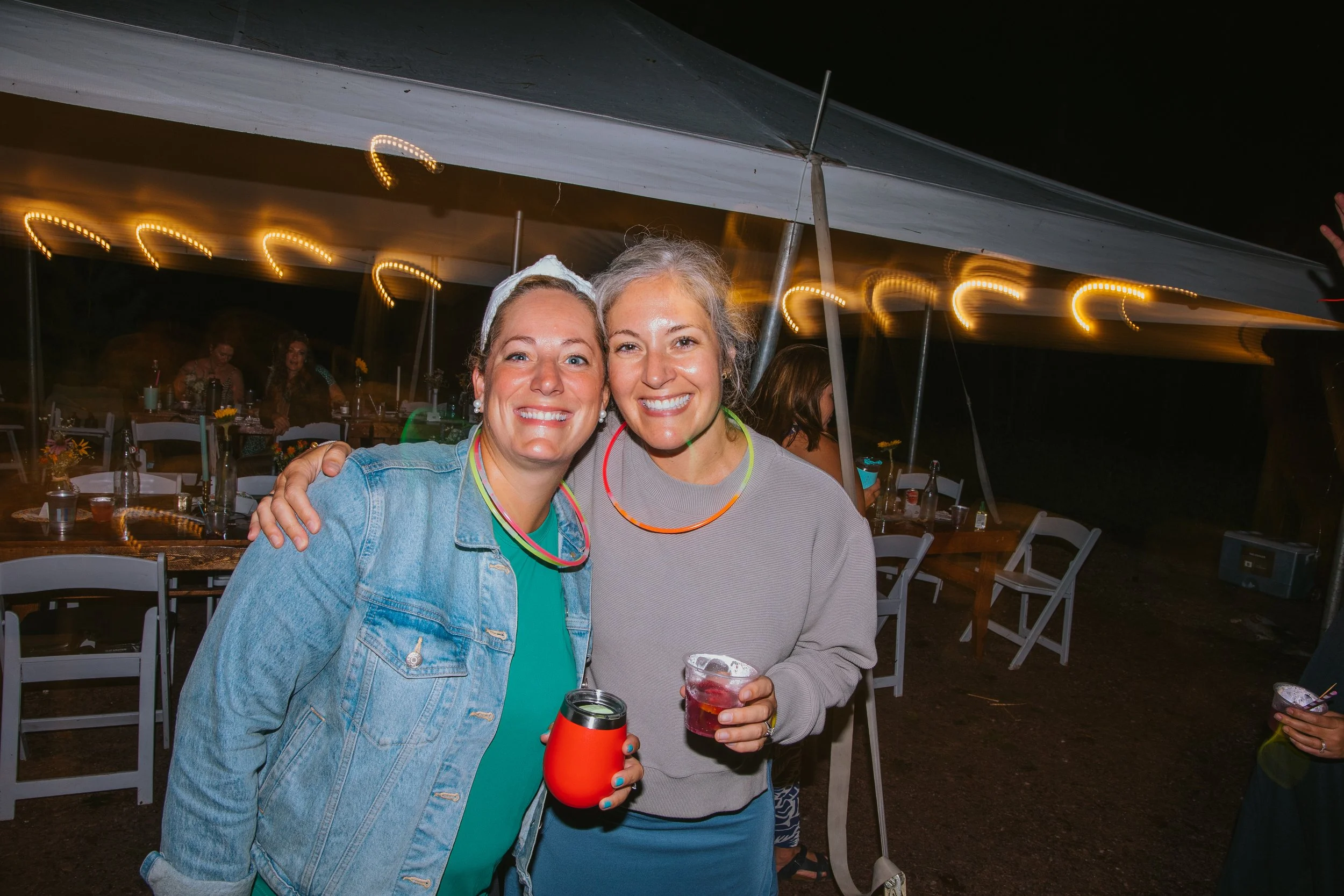 Two happy campers holding cocktails at Camp Lola Whiskey outdoor dinner under string lights in Wisconsin