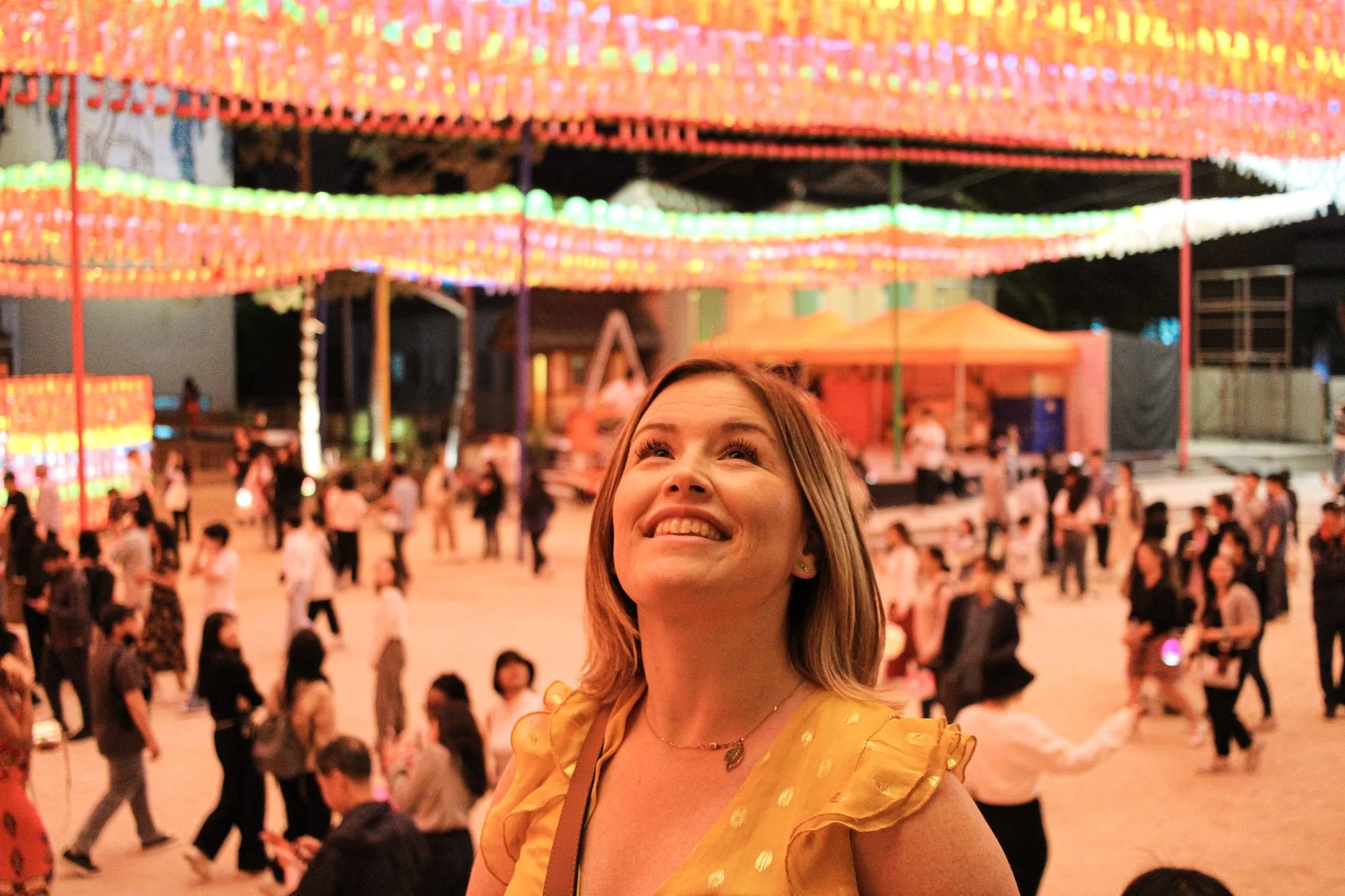 Woman in yellow polka dot dress smiling and looking up at vibrant overhead lantern display in crowded festival or market setting with warm lighting