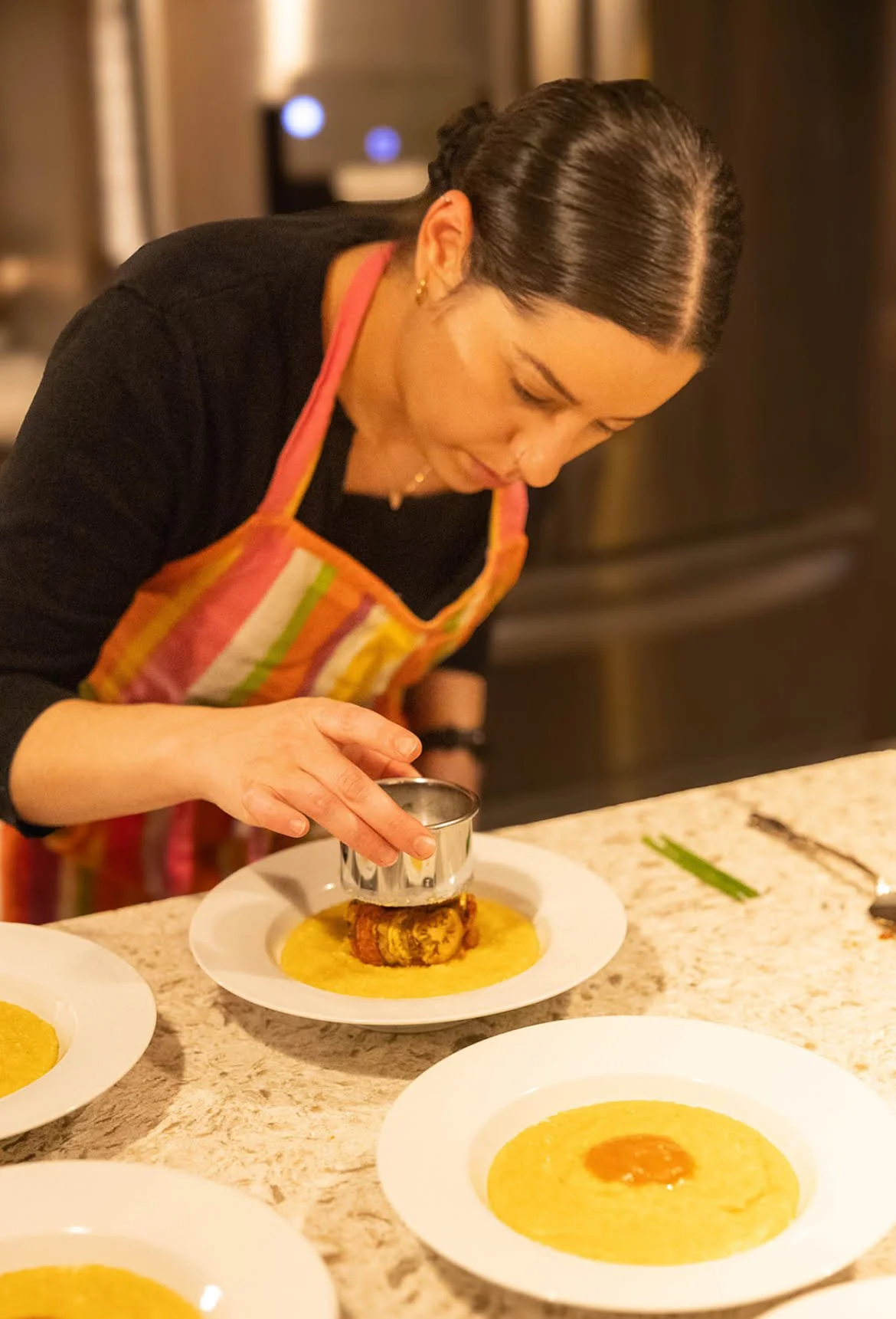 Here's an alt text option for this image: "Chef in colorful striped apron carefully plating a golden yellow sauce onto a white plate with a culinary mold, concentrating on presentation in a professional kitchen setting