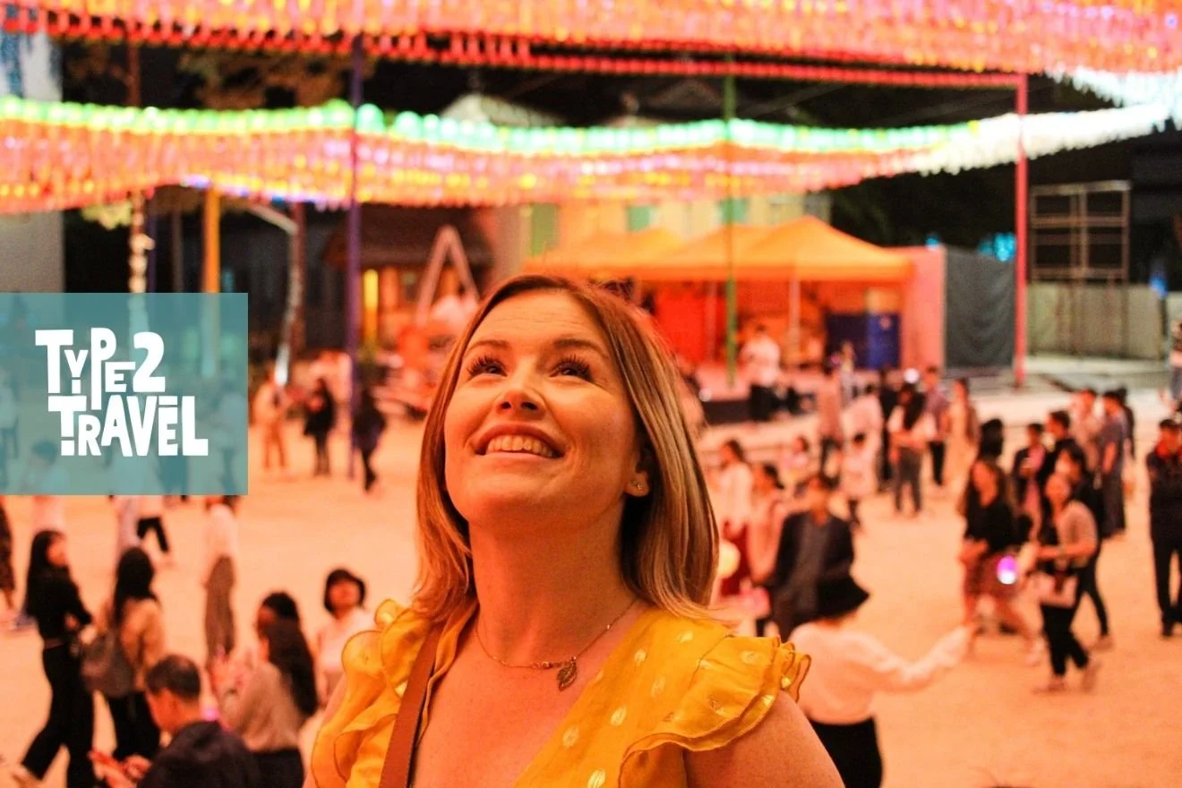 Kim Kephart looking up joyfully in a yellow ruffled dress at a vibrant outdoor ice skating rink with colorful lights and crowds in the background, Type 2 Travel podcast logo in corner