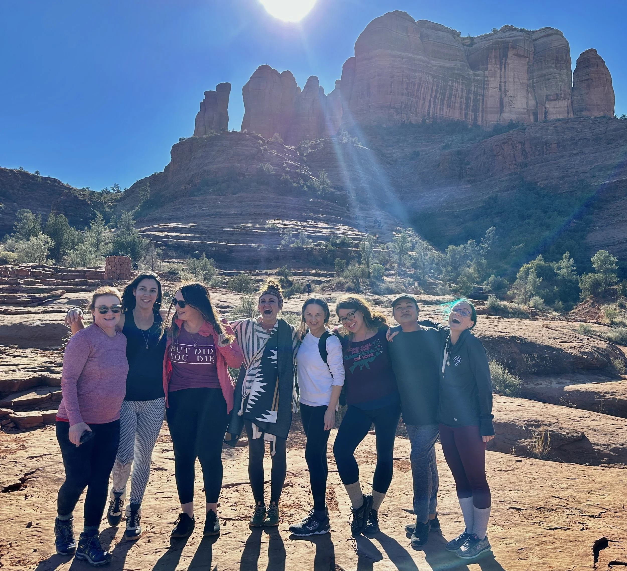 Group of nine women posing together outdoors in front of a large rocky mountain formation with clear blue sky and sunlight shining above.
