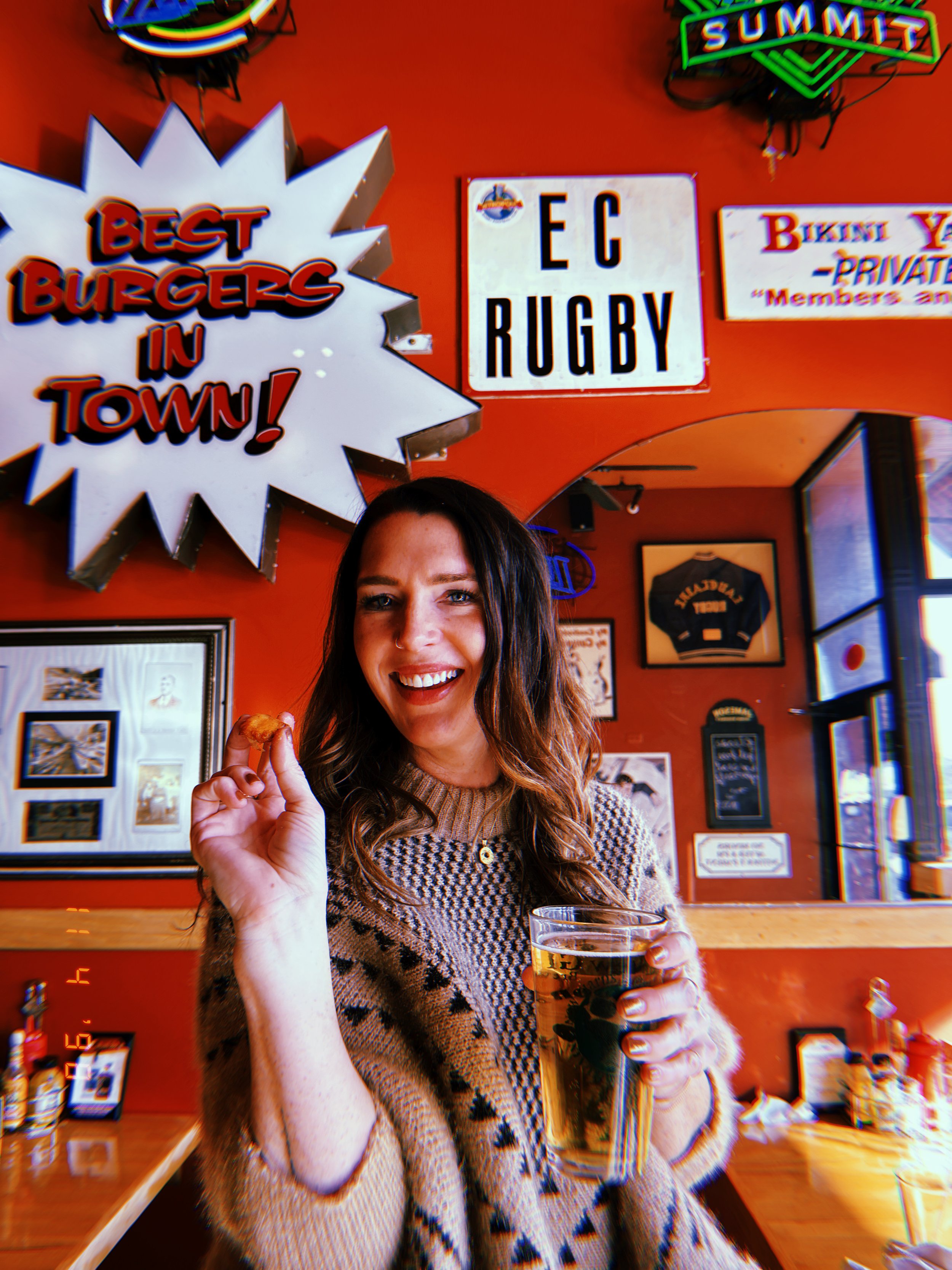 A smiling woman with long wavy hair holds a beer and a small food bite inside a lively bar decorated with bold red walls, neon signs, and memorabilia including an "EC Rugby" sign and a "Best Burgers in Town!" sign.