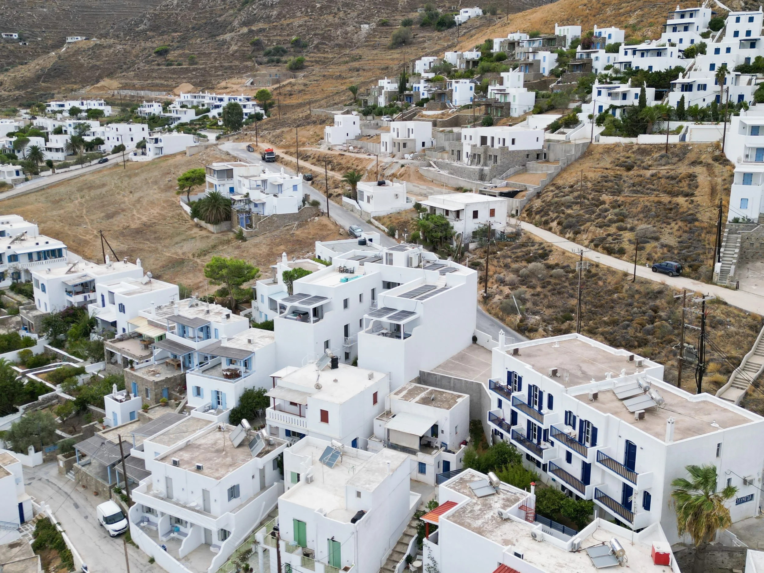 An aerial view of white Cycladic buildings nestled on a hillside, showing the characteristic architecture of Serifos island with its cubic houses, narrow streets, and the contrast between developed areas and the rugged landscape.