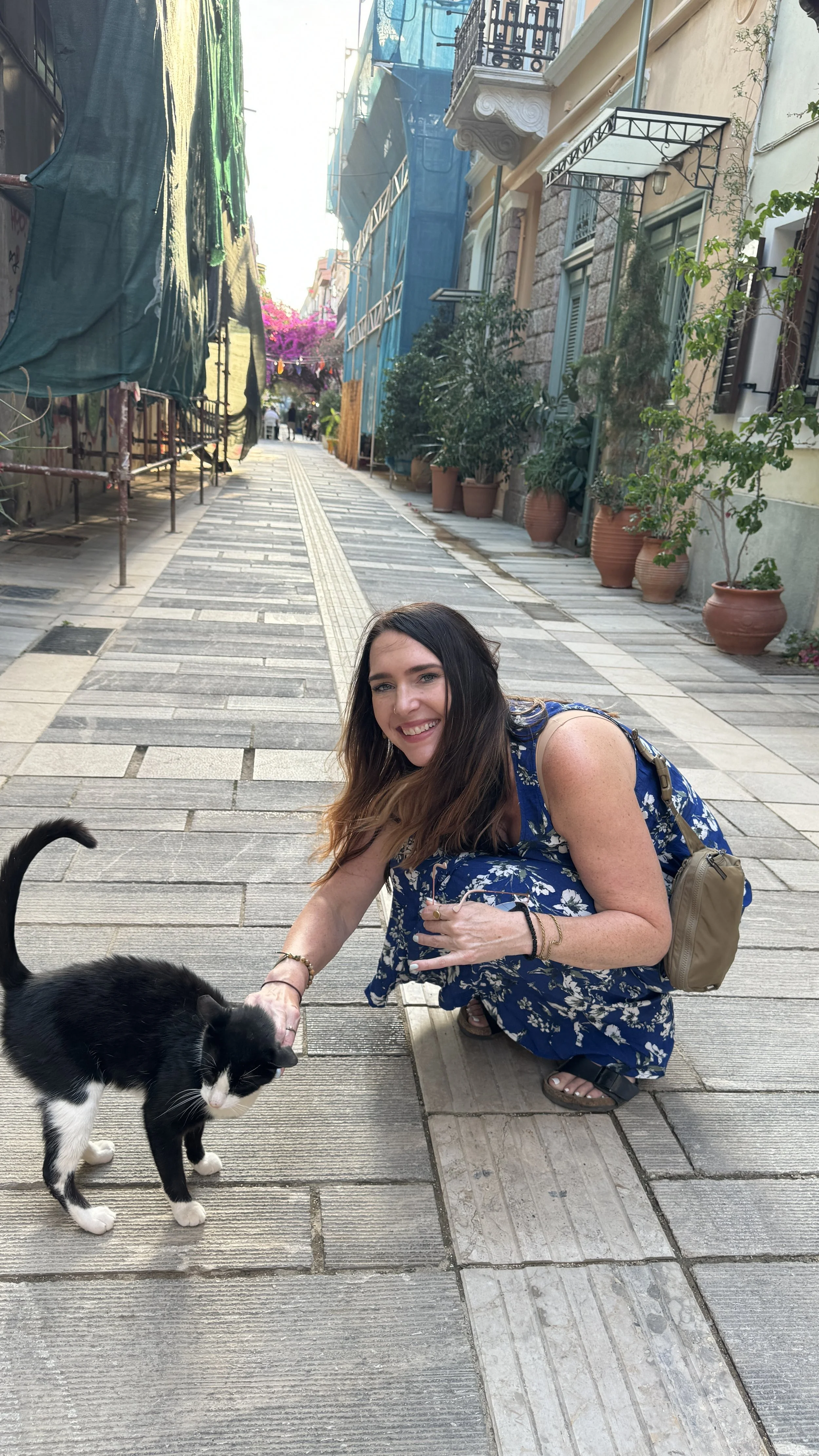 Laura Ericson crouching down to pet a black and white street cat in a cobblestone alley in Athens, Greece