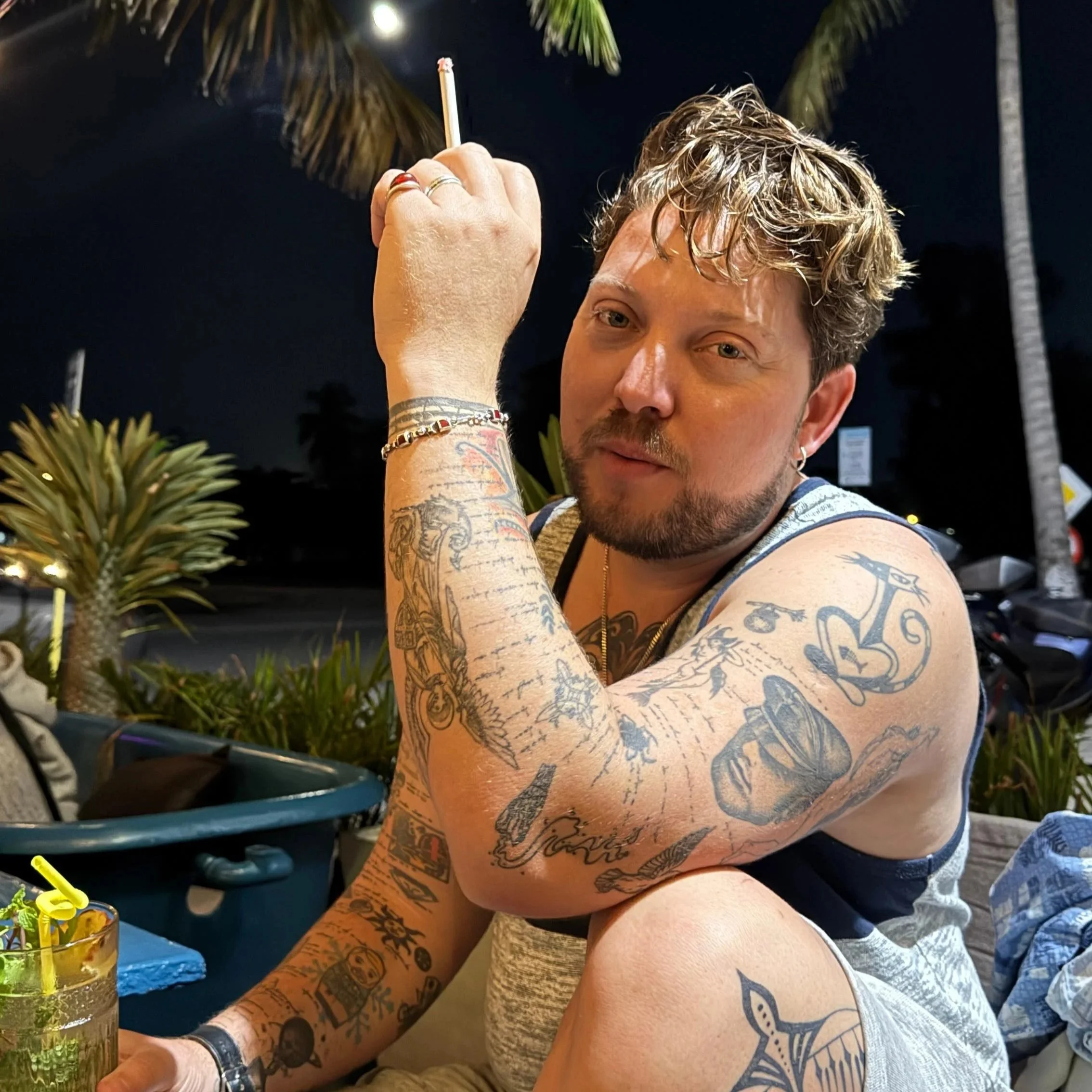 Person in tank top with tattoos relaxing poolside at night with tropical drink under palm trees and string lights
