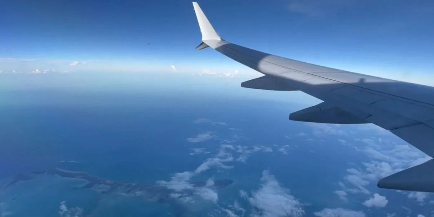 View from an airplane window showing the aircraft wing against a bright blue sky, with scattered clouds and a coastline below, where airplane etiquette starts at 35,000 feet.