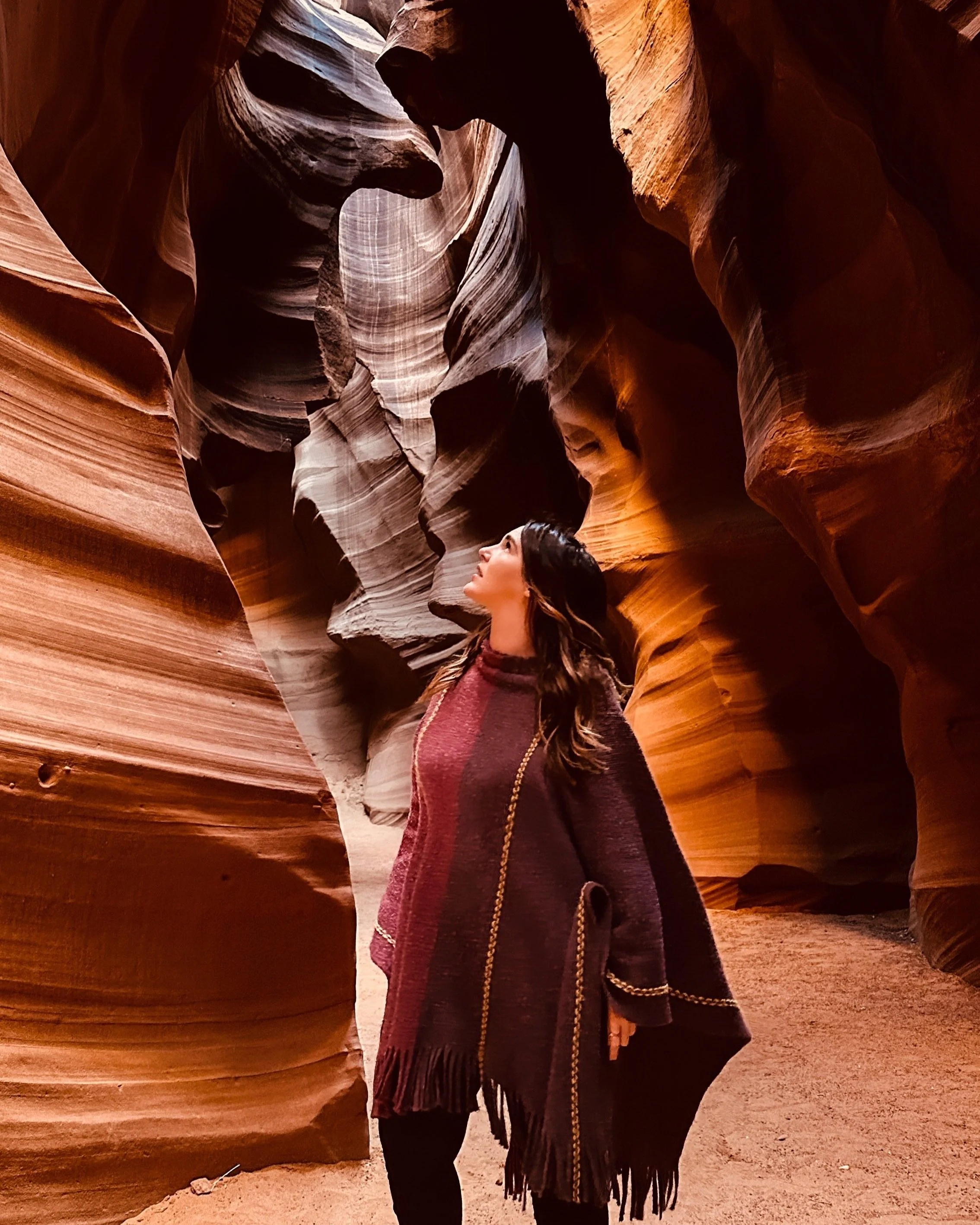 A woman standing inside a narrow slot canyon with smooth, layered sandstone walls in shades of orange, red, and brown, looking upward.