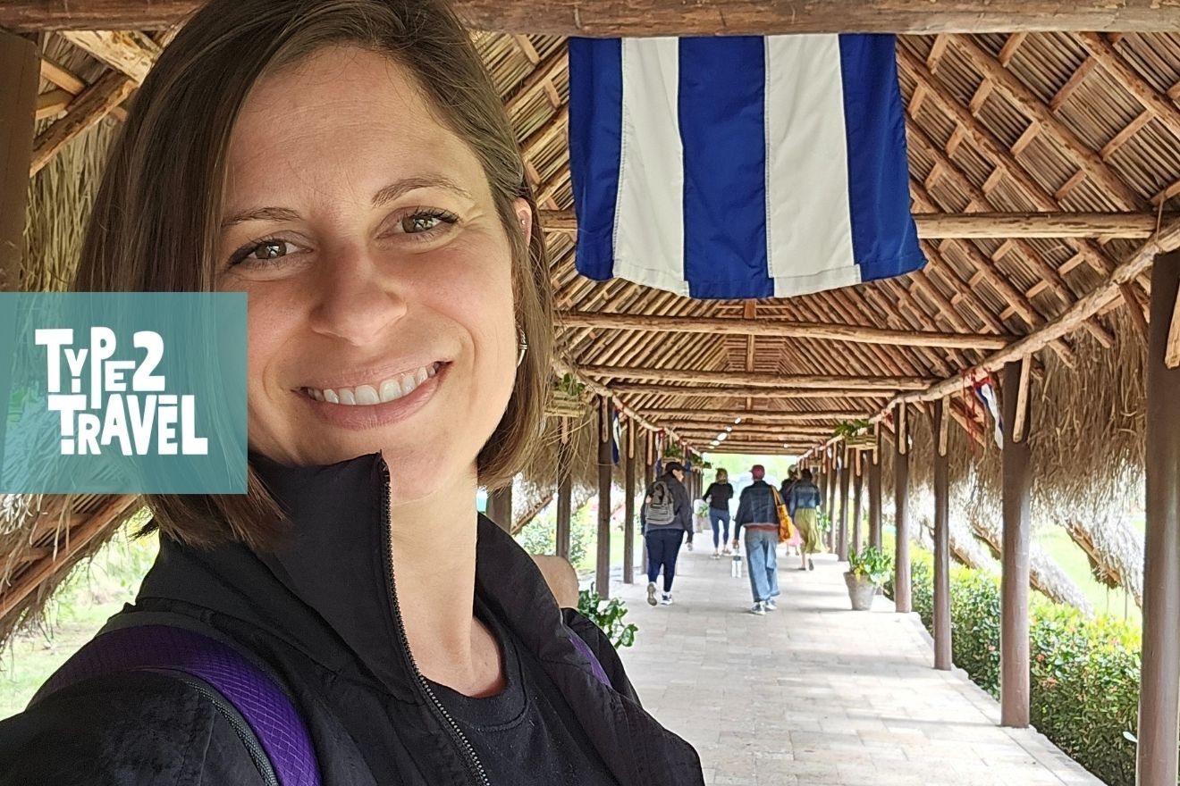 Type 2 Travel podcast guest Andrea at thatched roof walkway in Cuba with blue and white Cuban flag overhead