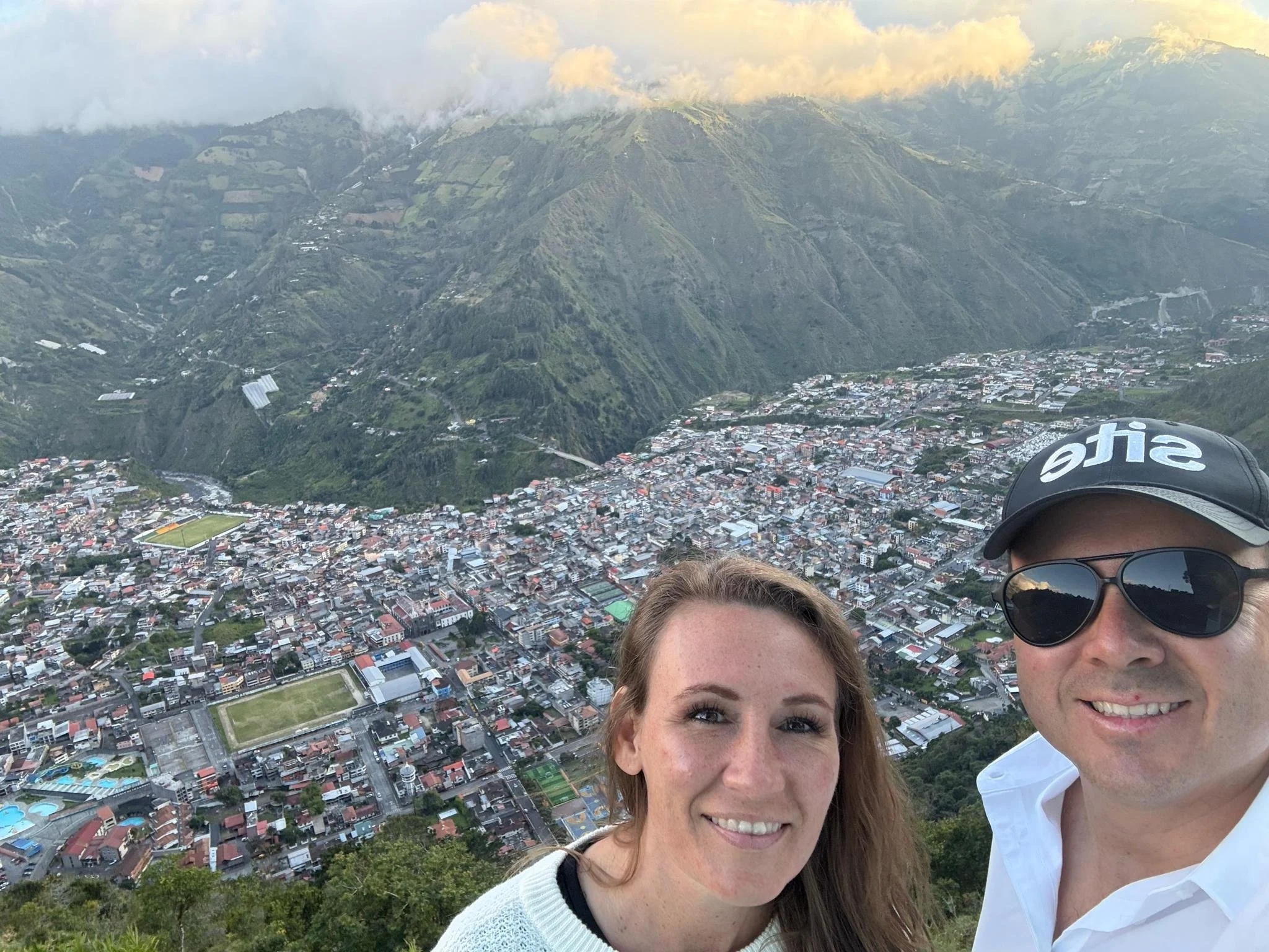 Two people smiling in a selfie overlooking the city of Baños nestled in a valley surrounded by Andean mountains in Ecuador