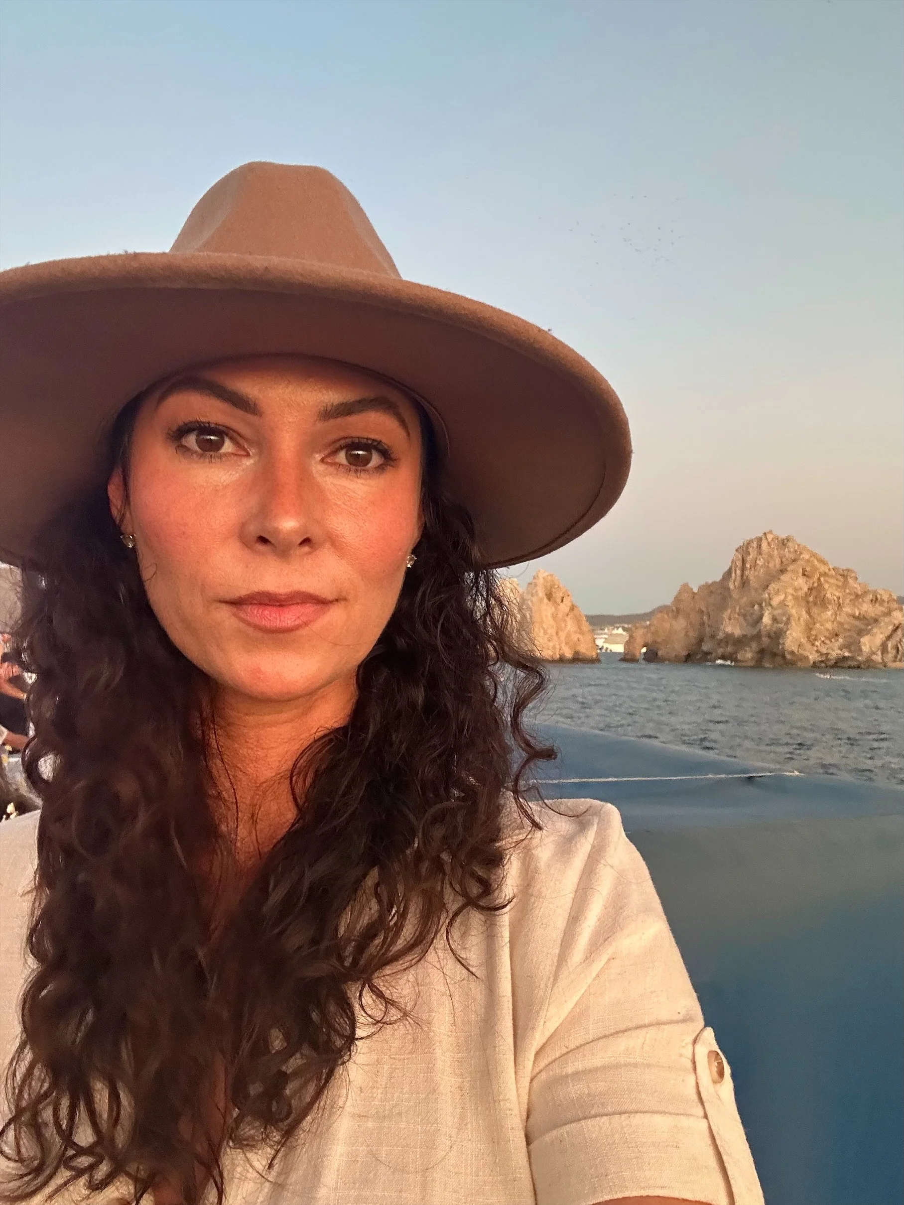 A woman with long, dark curly hair wearing a tan wide-brimmed hat and cream-colored top takes a selfie on a boat.