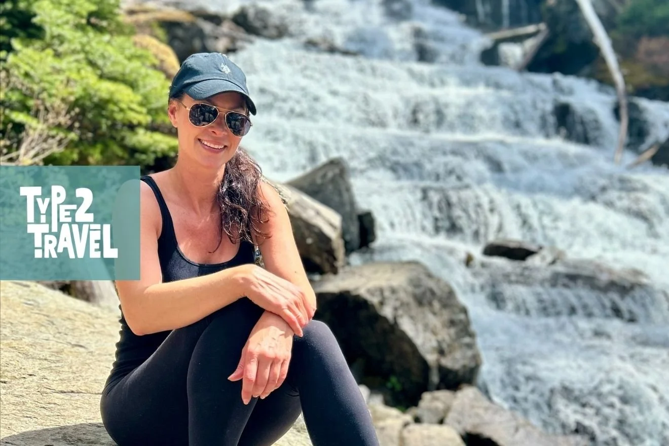 Tamara smiling in black athletic wear and sunglasses, sitting on rocks beside a cascading waterfall with Type 2 Travel podcast logo in corner