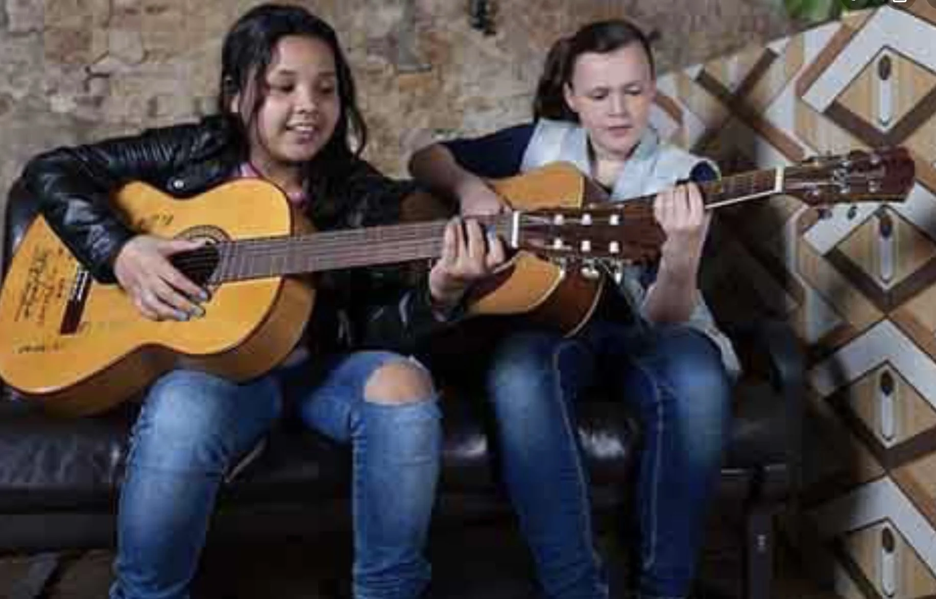 Two children, a girl and a boy, sitting on a bench and playing acoustic guitars.