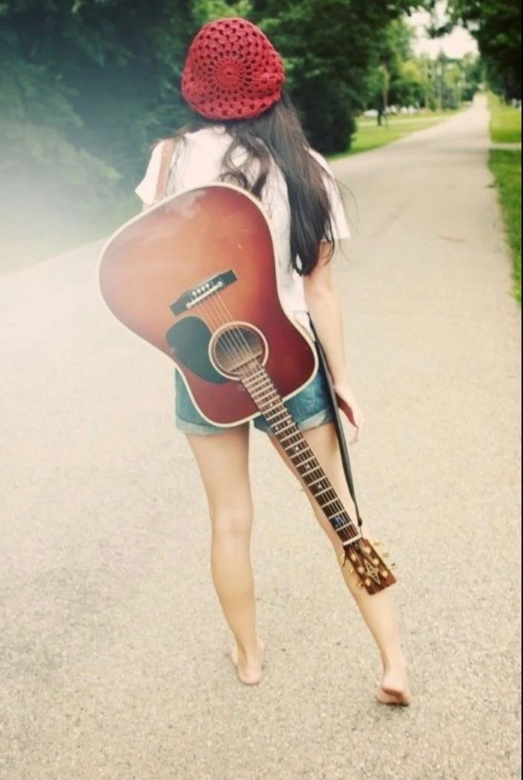 A young woman walking on a park path, carrying an acoustic guitar on her back, wearing a red crocheted hat, a white t-shirt, and denim shorts, with trees and greenery lining the path.