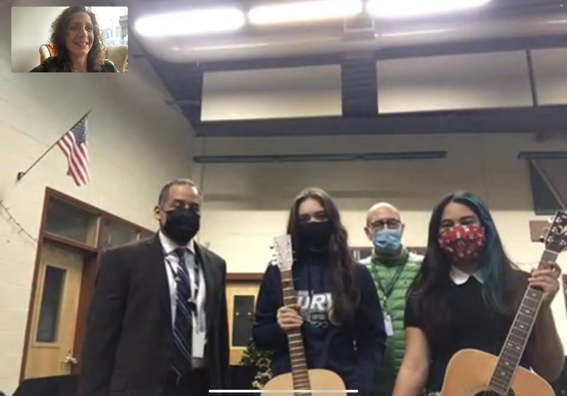 Four people wearing masks holding guitars in a room with an American flag, Christmas decorations, and a beige brick wall.