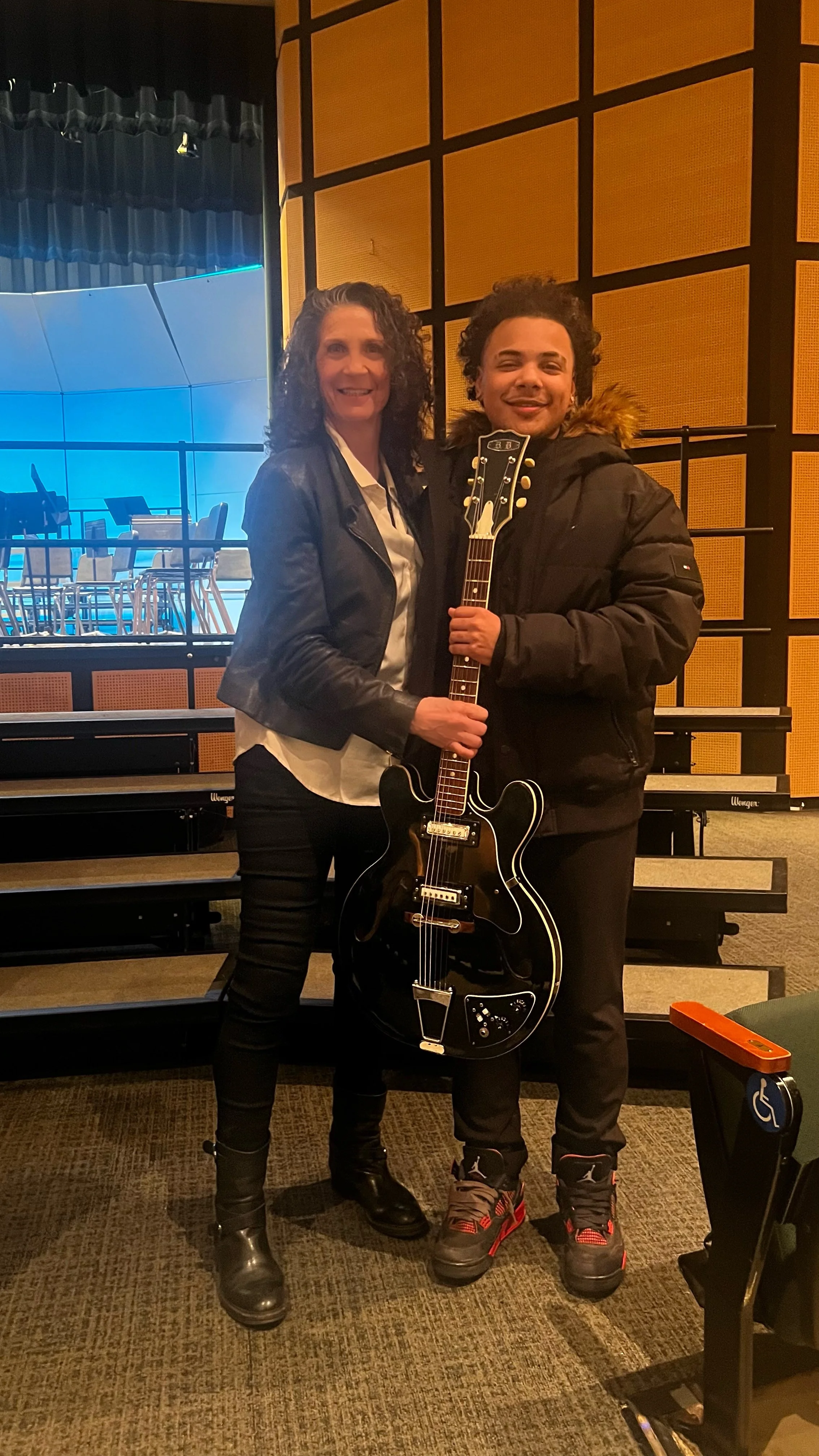Two women standing on a stage in a music venue, holding a black electric guitar. The woman on the left is wearing a leather jacket, white shirt, and black pants, while the woman on the right is dressed in a black coat with a fur-lined hood and black 