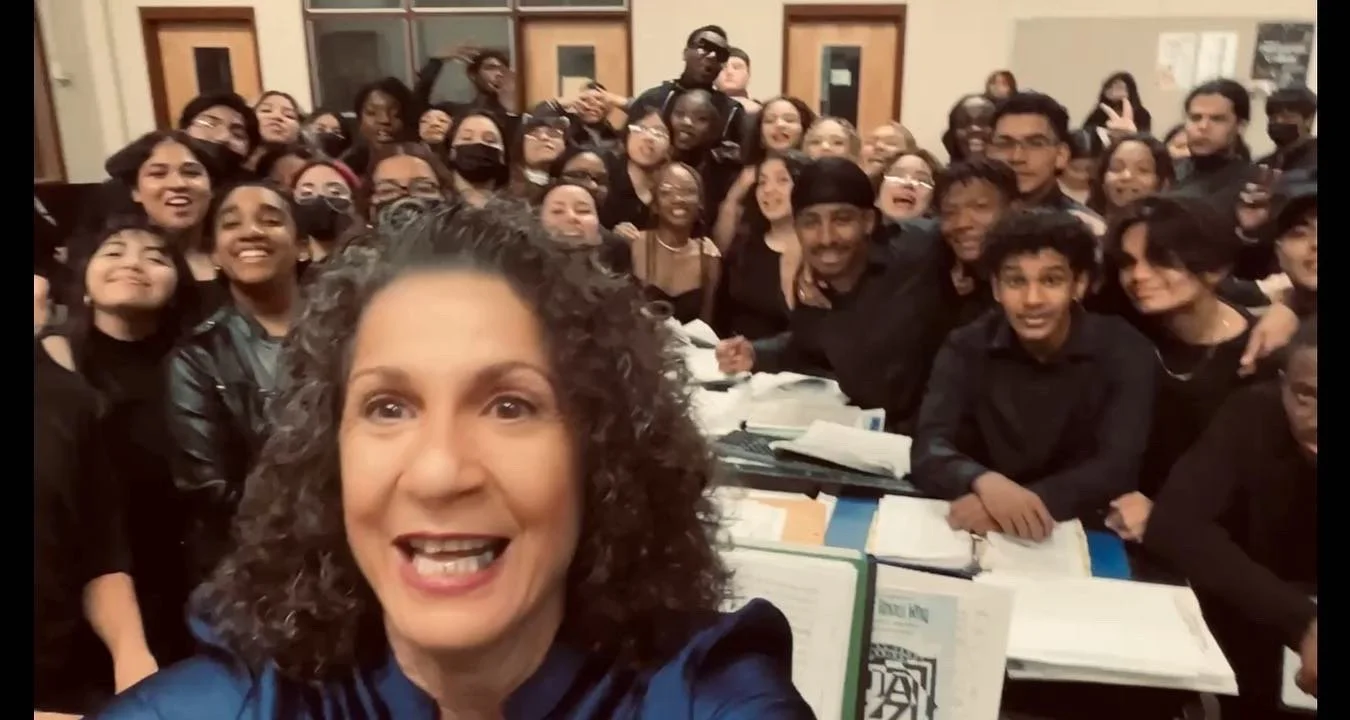 A woman taking a selfie with a large group of smiling and excited diverse students or young people, some wearing masks, gathered behind her in a classroom or auditorium setting.