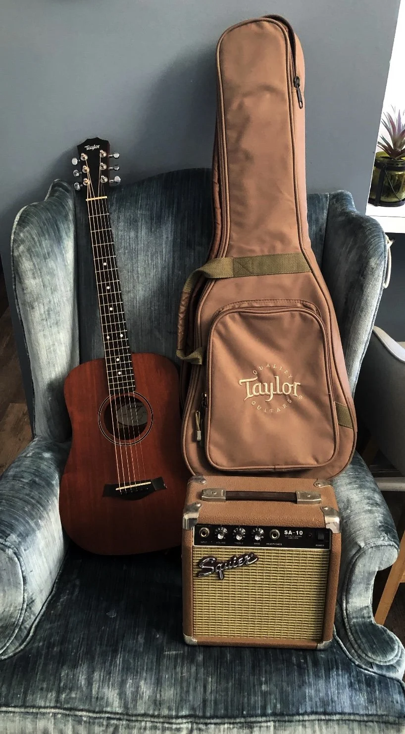 An acoustic guitar, a Taylor gig bag, and a small guitar amplifier on an upholstered armchair.