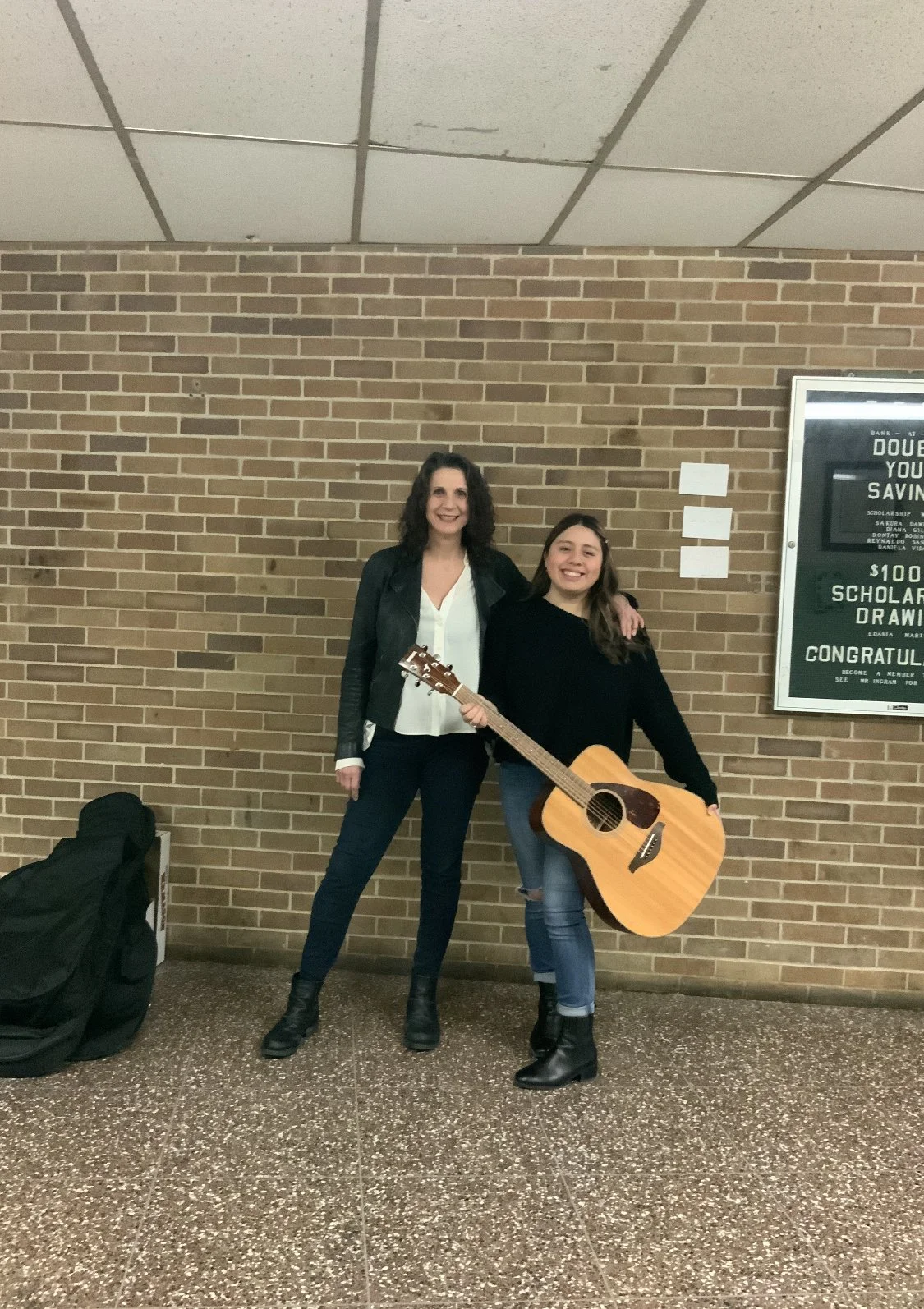 Two women, one holding an acoustic guitar, standing against a brick wall in a hall or corridor.
