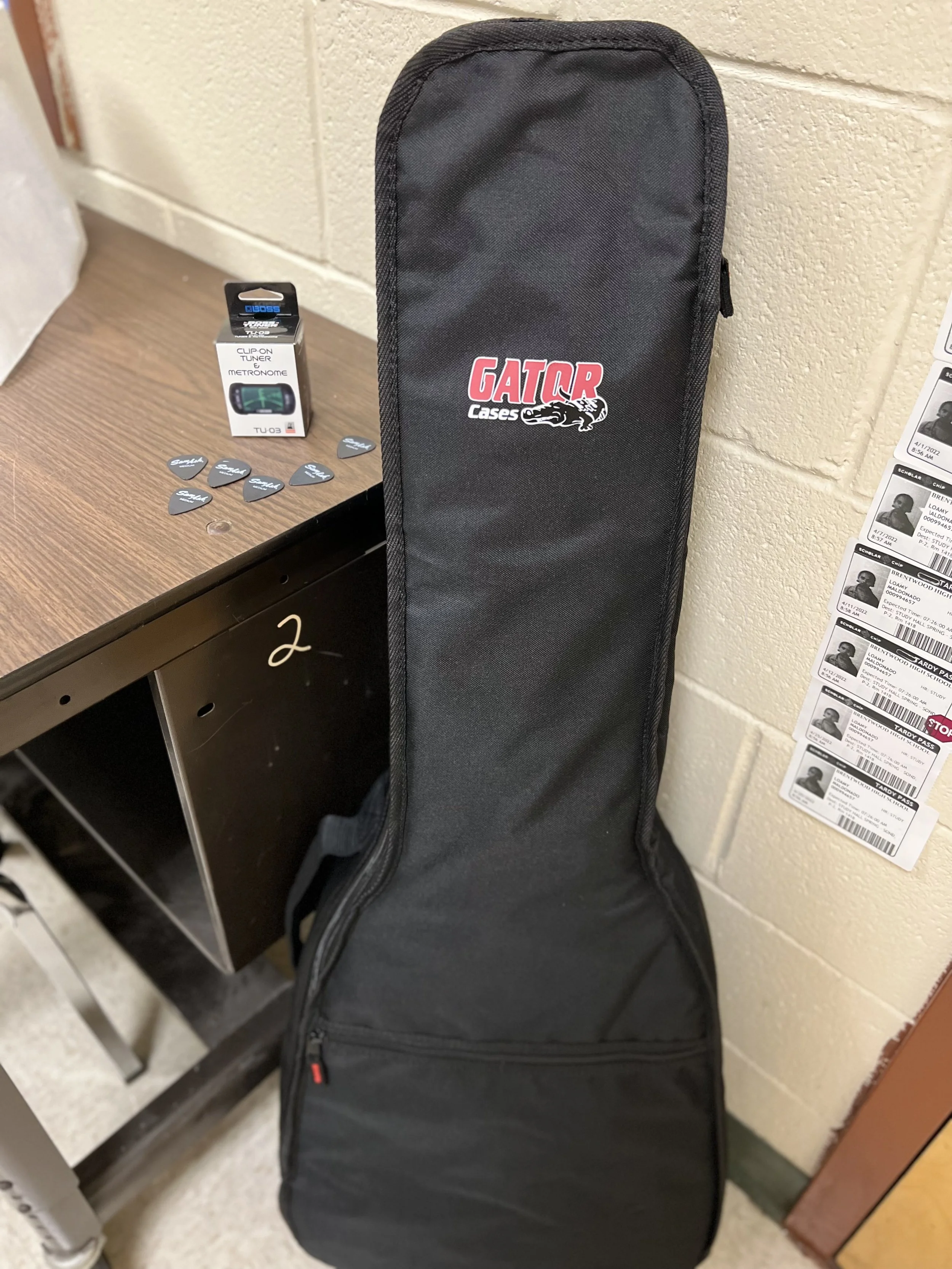A black Gator Cases guitar gig bag leaning against a beige brick wall in a room with label and ticket stubs on a wall.