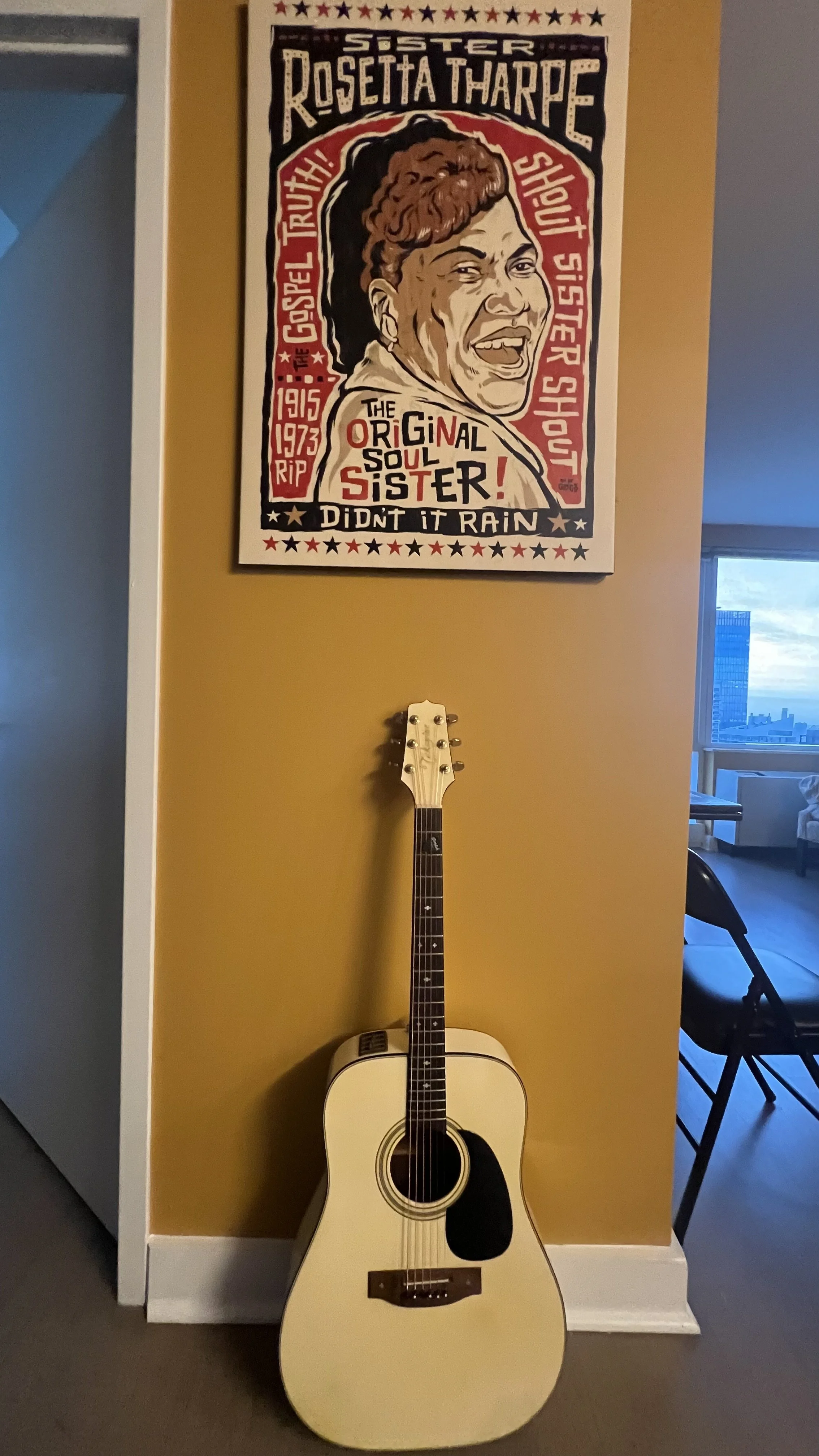 An acoustic guitar leaning against a yellow wall below a colorful poster featuring Sister Rosetta Tharpe, with artistic text and a portrait of her.