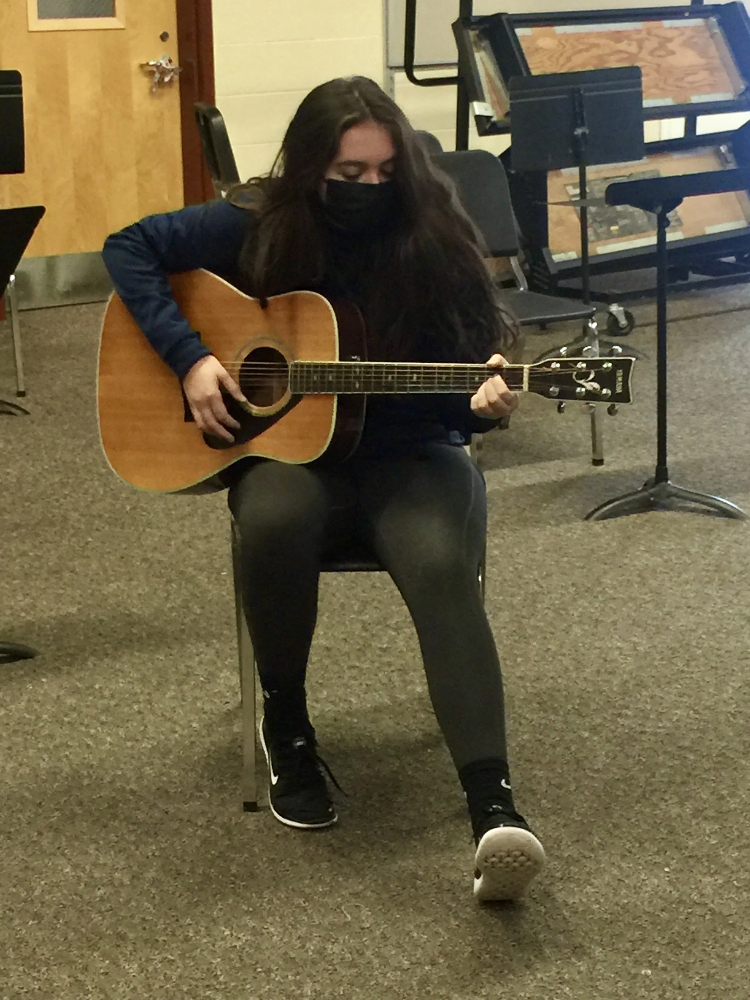 A young woman with long dark hair, wearing a black face mask, black leggings, a dark jacket, and black sneakers, sitting on a chair and playing an acoustic guitar in a room with musical equipment and a brown carpeted floor.