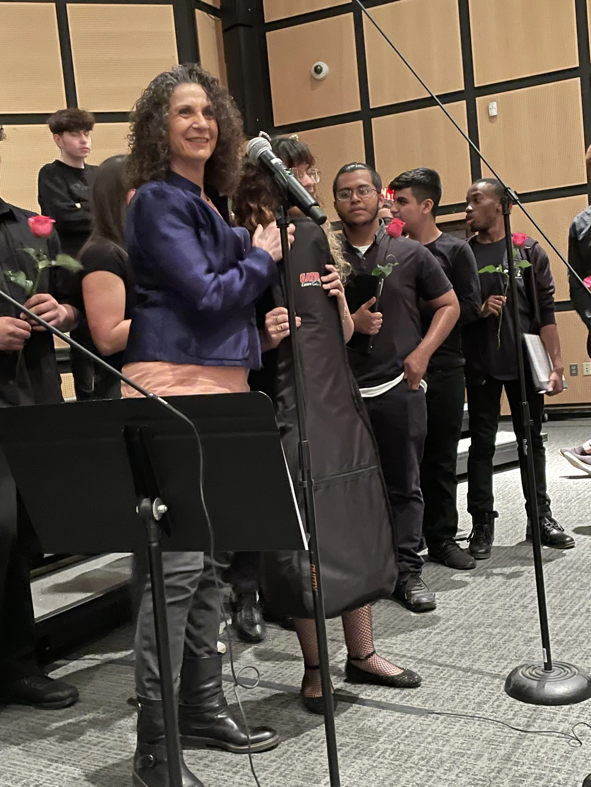 A woman speaking at a microphone with students holding roses behind her in a classroom or auditorium setting.
