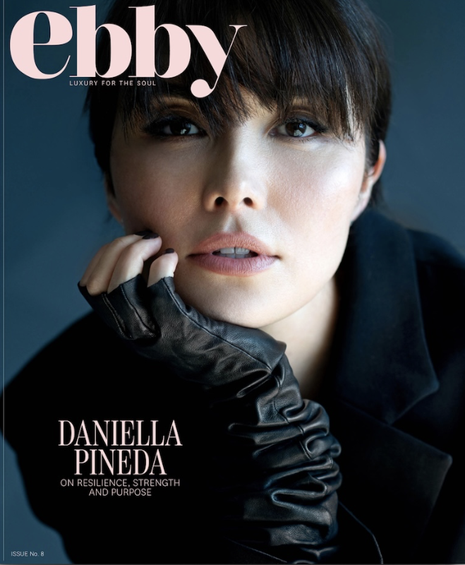 Close-up of a woman with short dark hair and striking eyes, wearing a black glove on her left hand and a black jacket, on the cover of Ebony magazine. The magazine features Daniella Pineda, focusing on resilience, strength, and purpose.