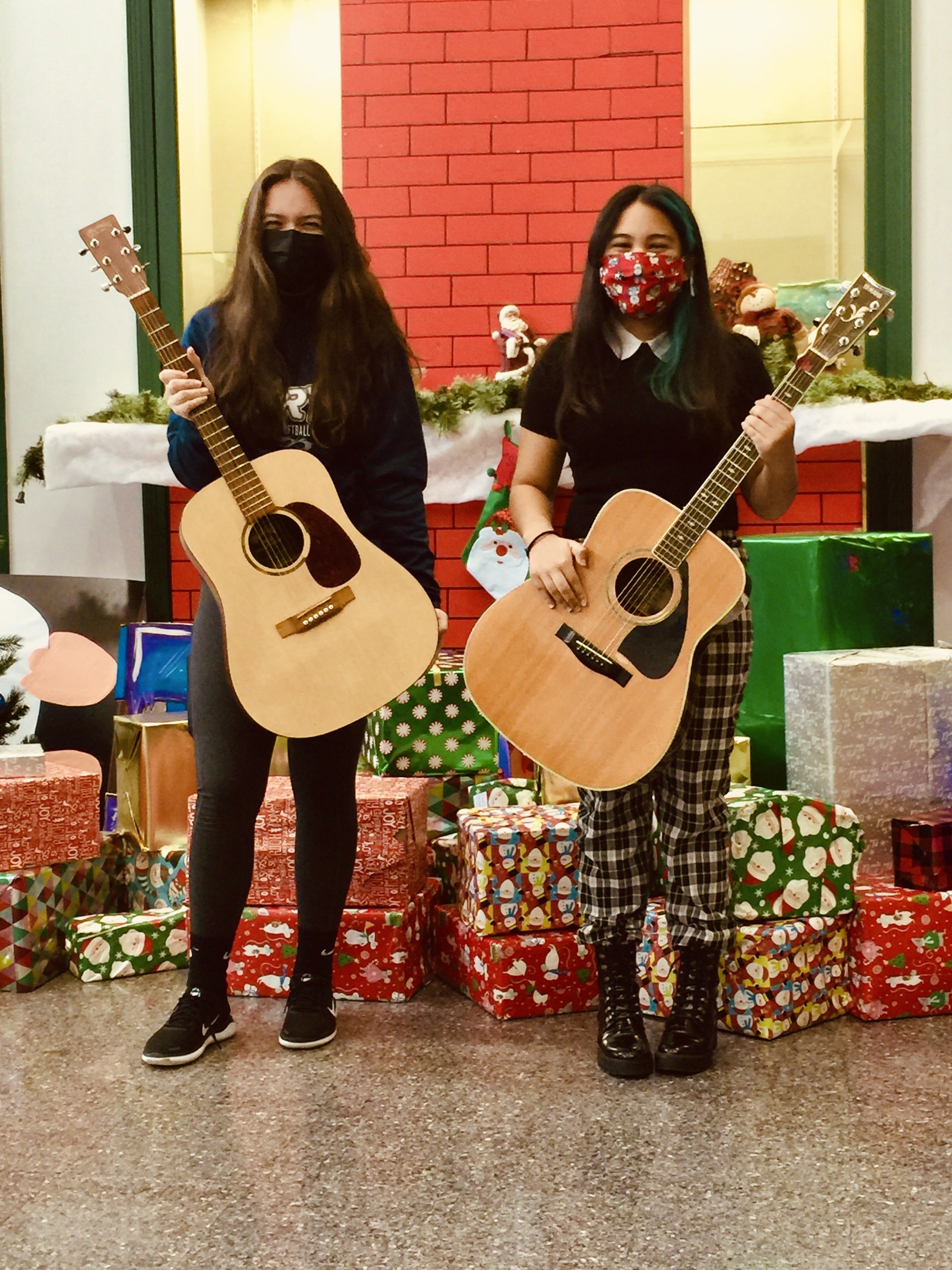 Two young women standing in front of Christmas decorations, each holding an acoustic guitar, wearing masks, and surrounded by wrapped gifts.