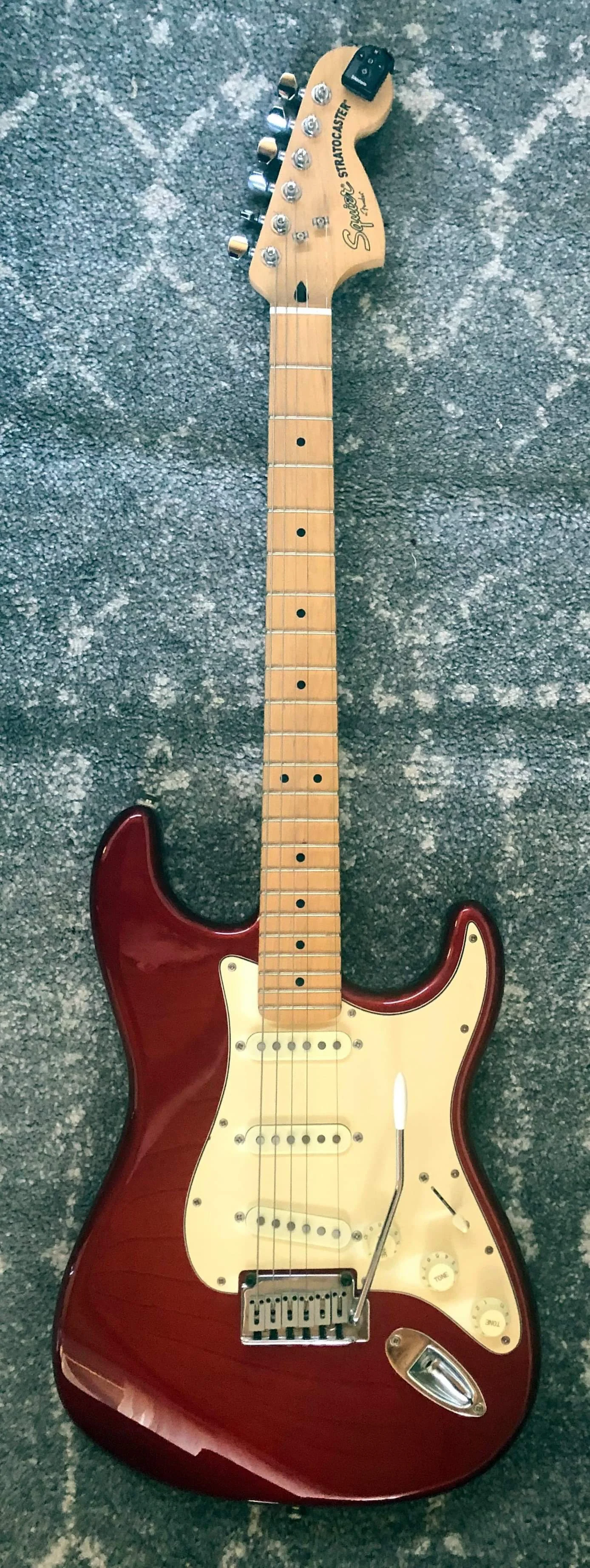 Electric guitar with a red body, cream pickguard, maple neck and fretboard, six pickups, and a tremolo arm, lying on a gray carpet.