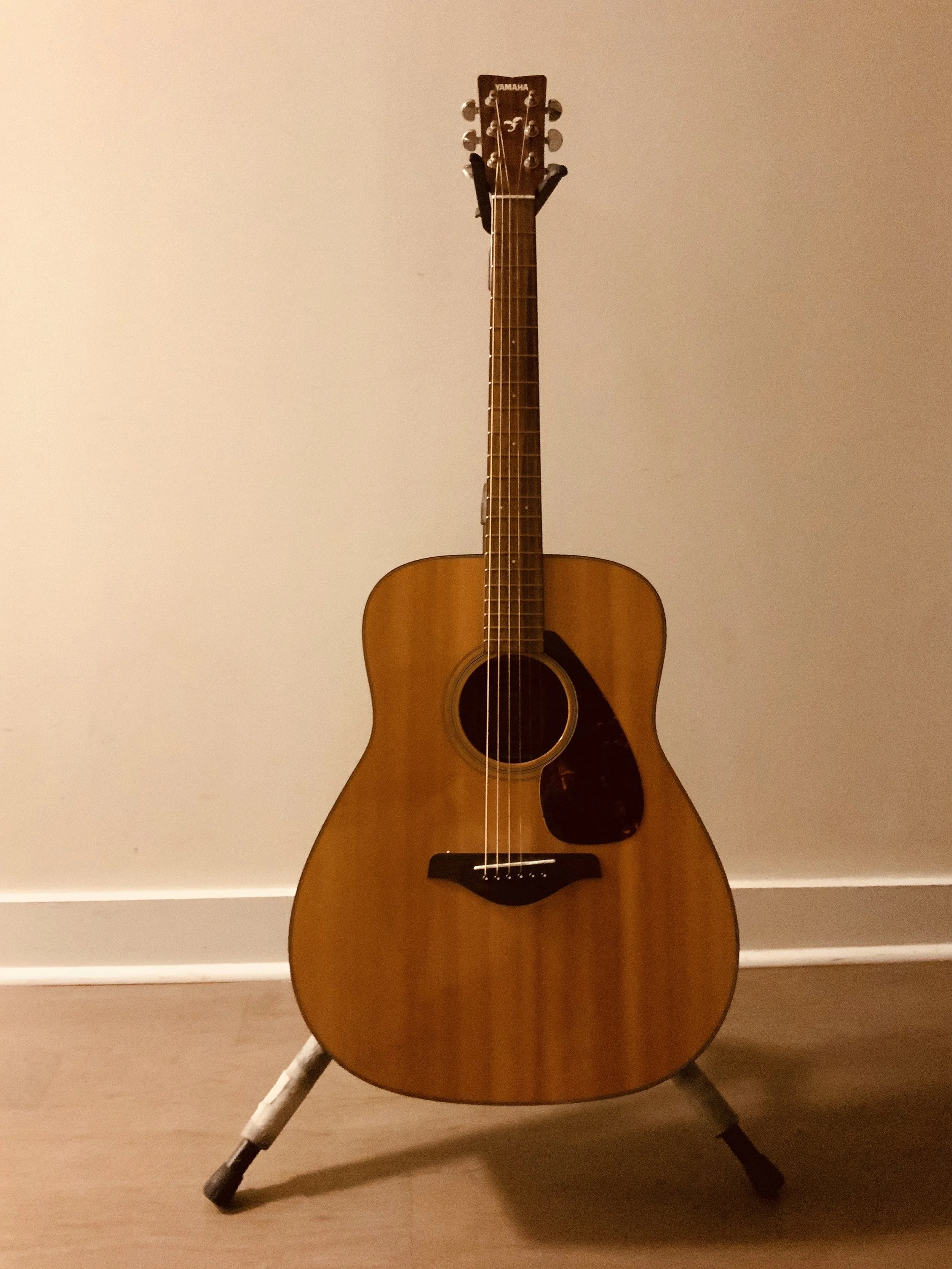 An acoustic guitar standing on a wooden floor against a plain wall, with a dark brown pickguard and light-colored wood body.