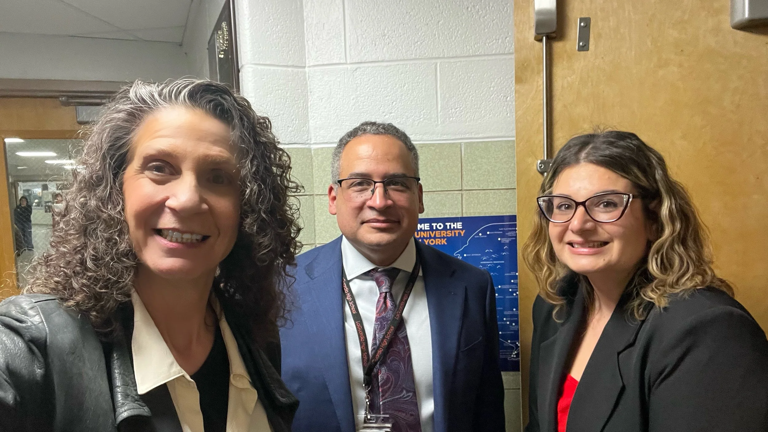 Three people, two women and one man, standing close together smiling for a photo indoors near a door and a school sign.