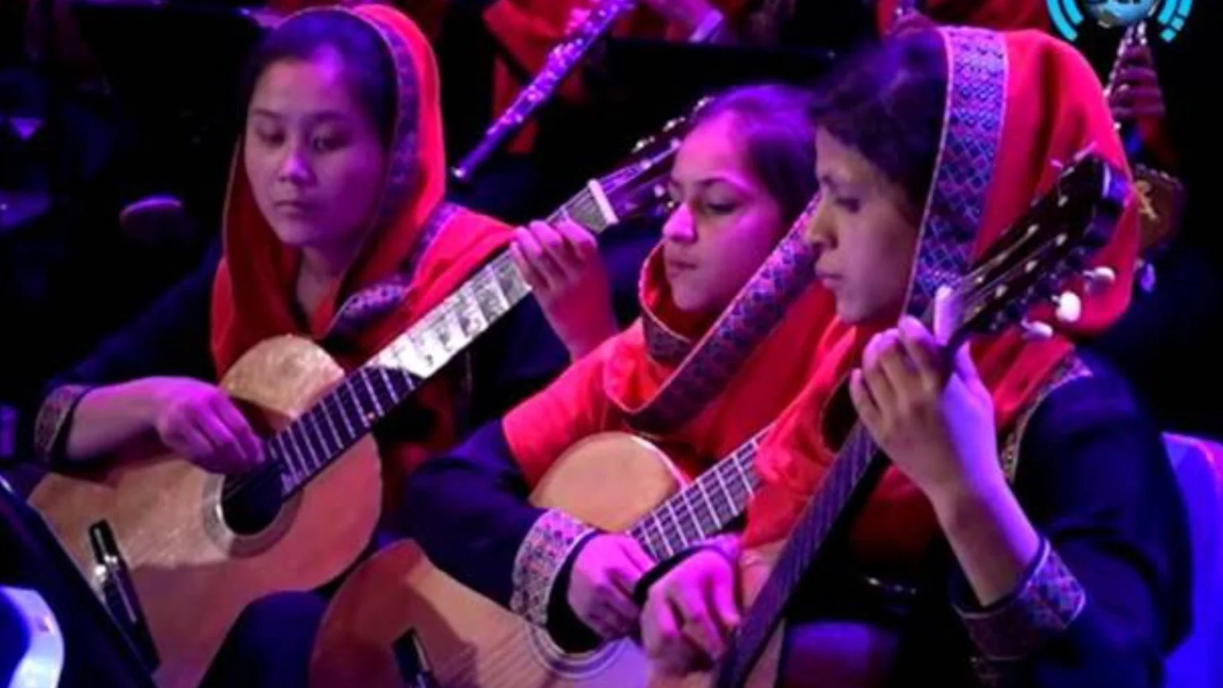 Three women in red and black traditional attire playing acoustic guitars during a performance.