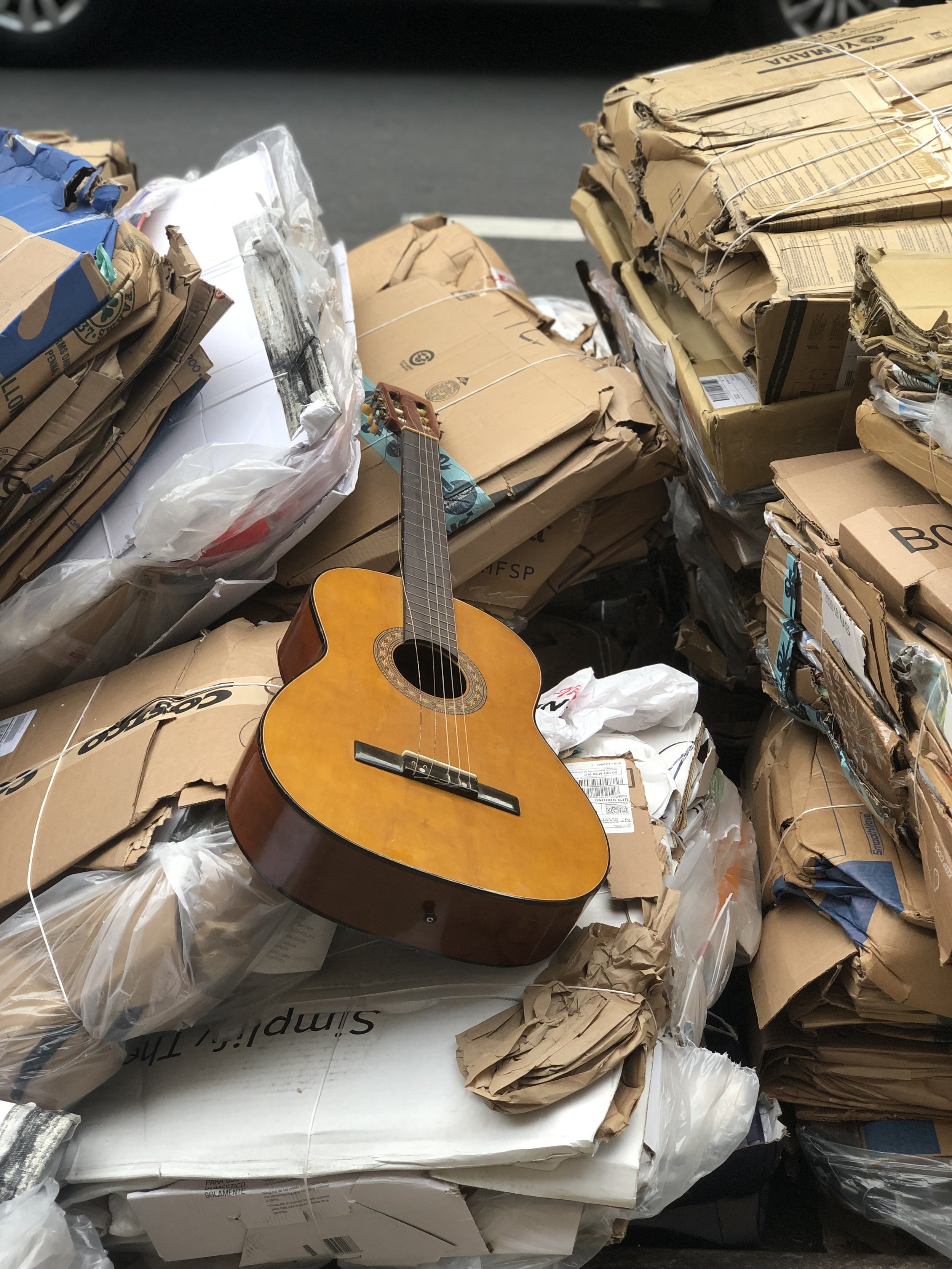 An acoustic guitar lying on a pile of flattened cardboard boxes and packaging materials in an outdoor setting.