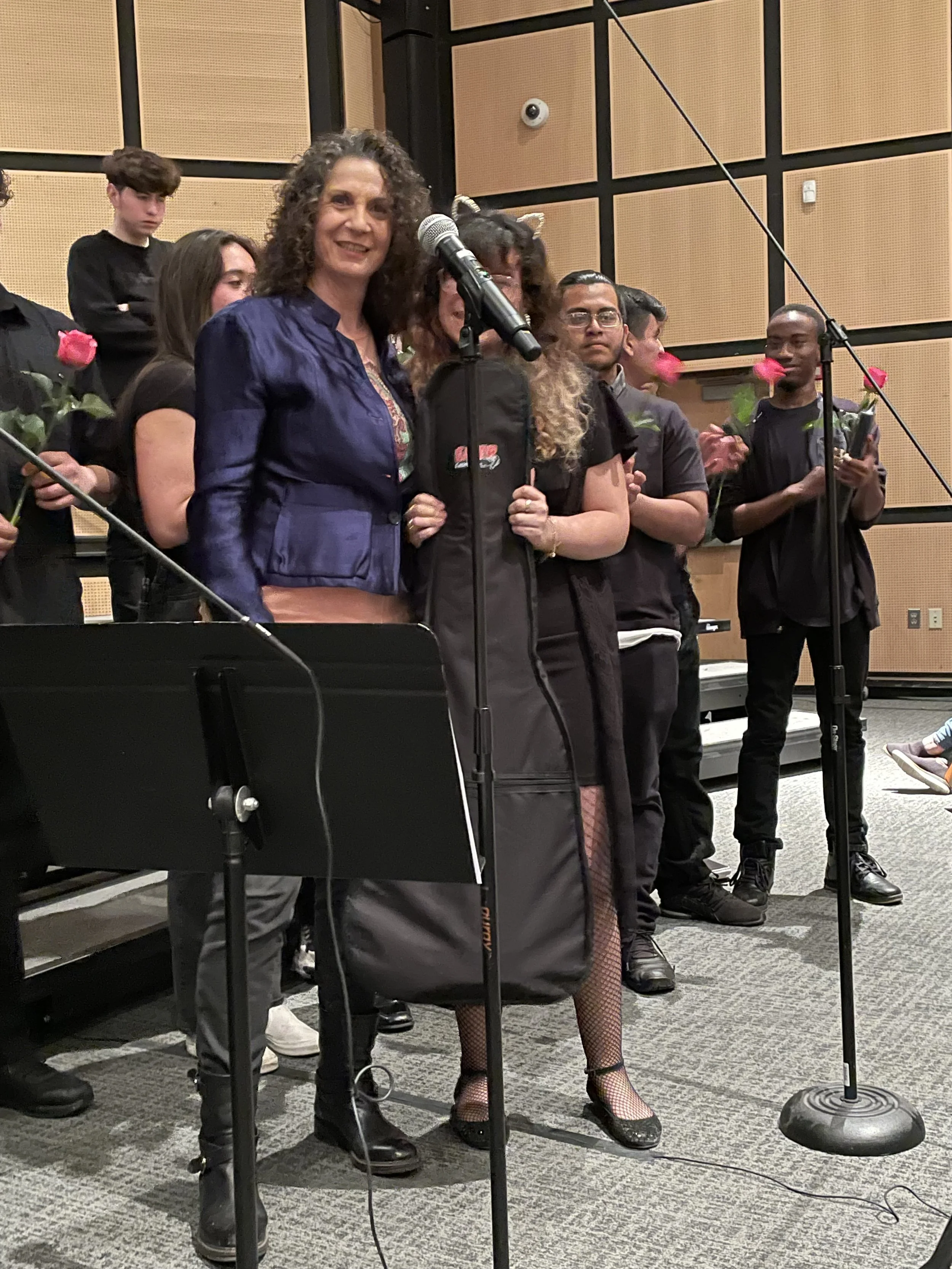 A group of people on stage at a public event, with a woman in a blue jacket speaking into a microphone, surrounded by others holding roses and certificates.